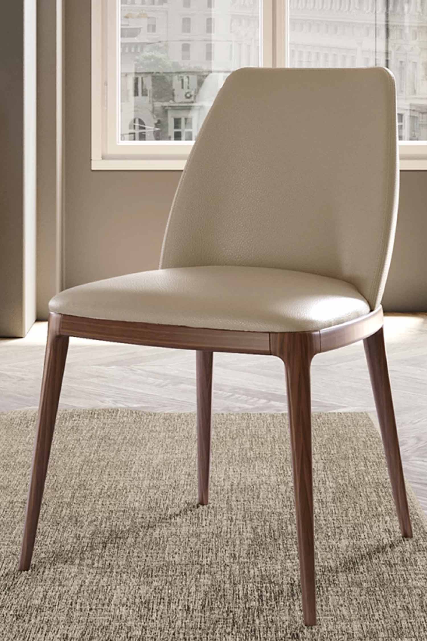 Close-up, vertical shot of the Max luxury dining chair featuring light beige textured leather upholstery and a warm brown wooden frame on a textured rug near a window.