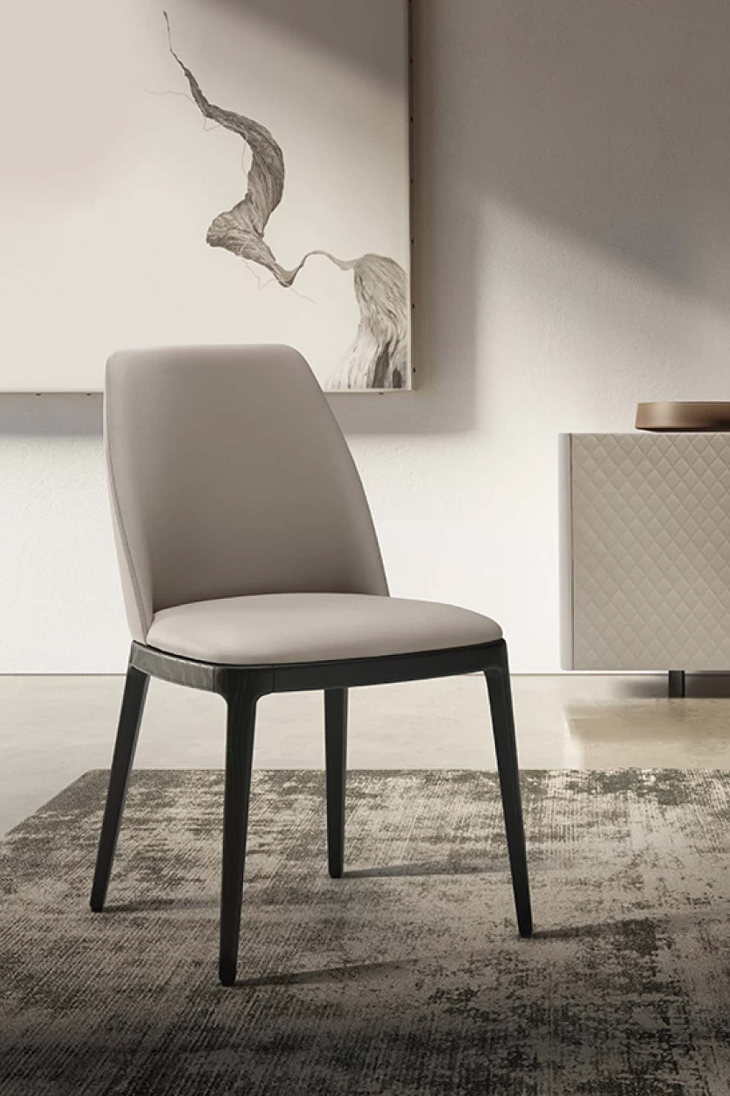Close-up of the Max luxury armless dining chair with light taupe leather upholstery and a dark wood frame, placed on an abstract rug in front of a modern sideboard and art.