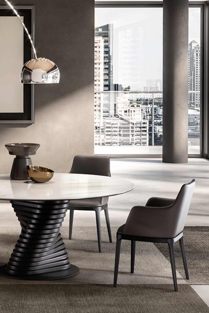 Two dark gray Max armchairs surrounding a round white marble dining table with a sculpted black spiral base, set in a modern high-rise dining space with a large window view.