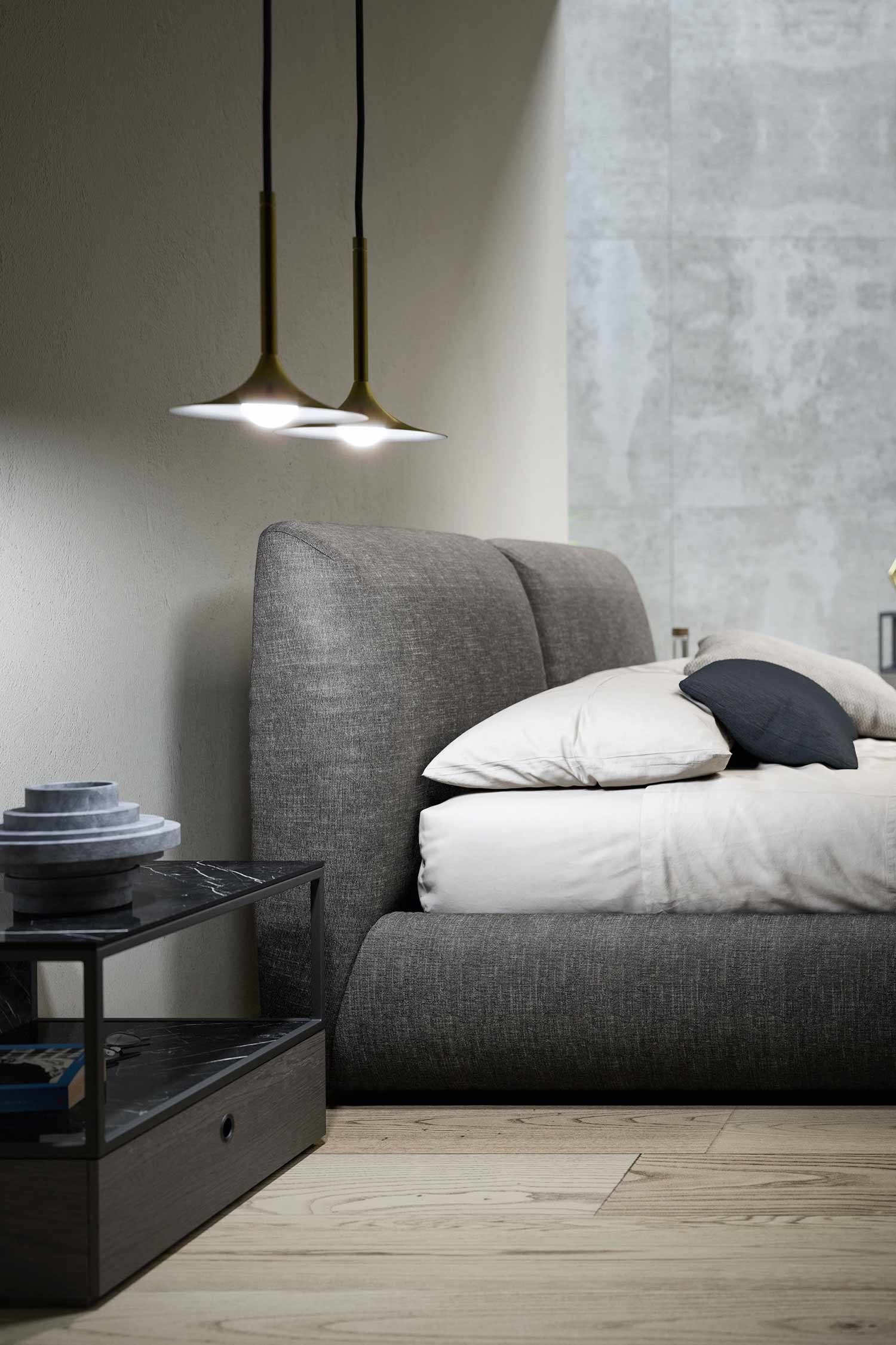 Close-up detail of the dark gray upholstered bed's split headboard and white and navy bedding. To the left, a modern nightstand with a dark marble-look top is visible, and two brass pendant lights with circular shades hang above. The background features light wood flooring and a gray textured wall.