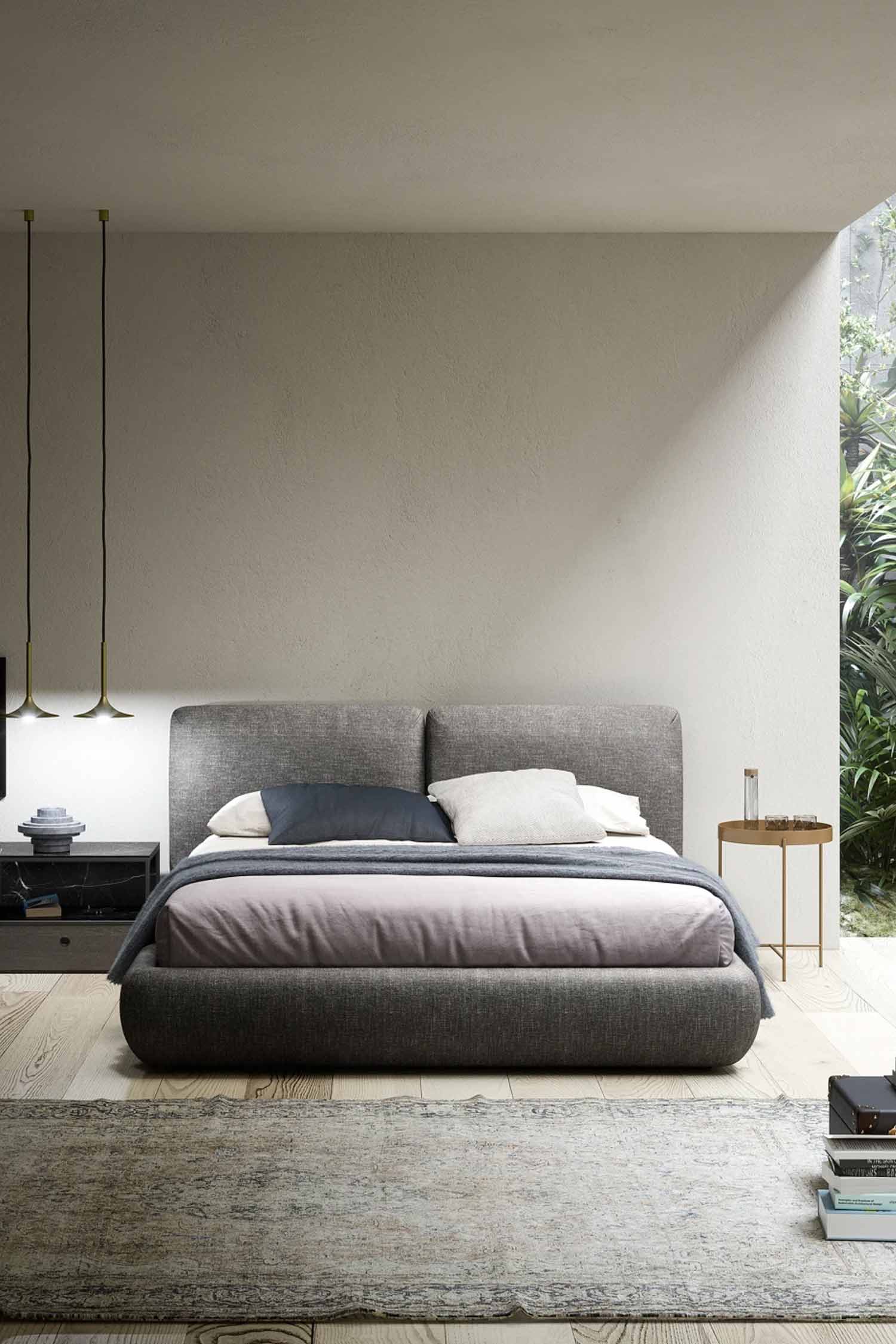 A full-view shot of a modern bedroom featuring a dark gray upholstered bed with a low, rounded profile and split headboard. The bedding includes a light mauve duvet, gray throw, and neutral pillows. The room has light, textured walls, minimalist pendant lights above a dark nightstand on the left, and a small copper-toned side table on the right. A lush outdoor garden is visible through an open wall on the right.