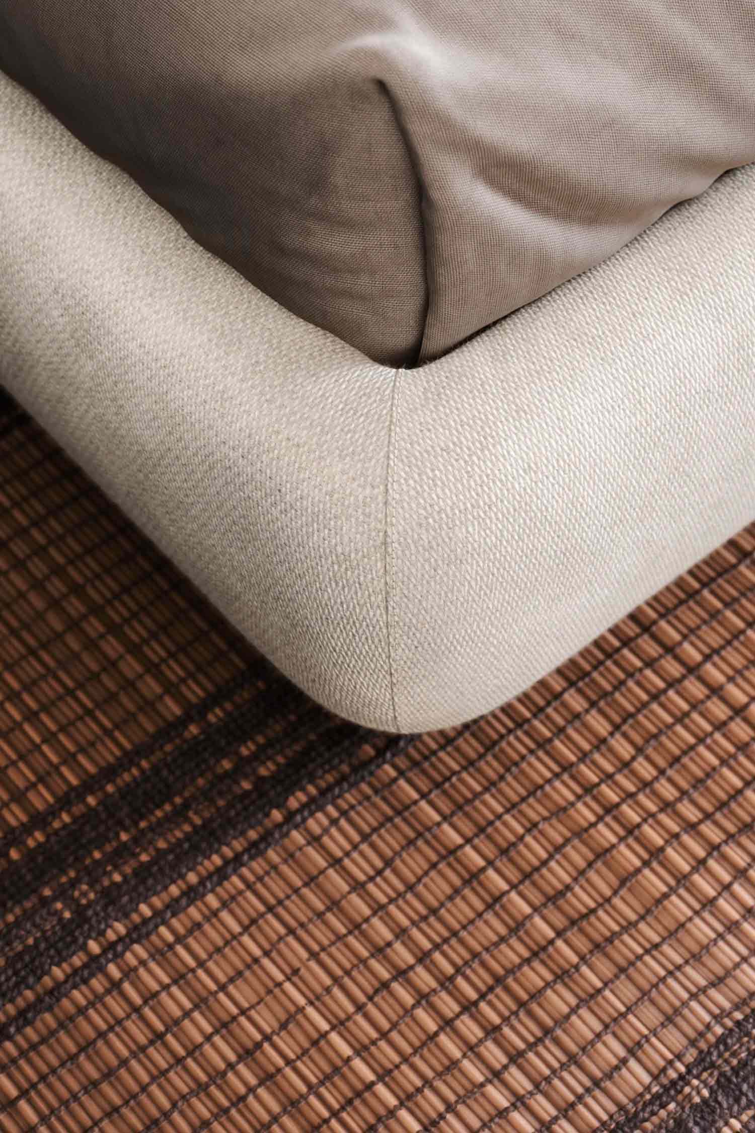 Extreme close-up of the rounded corner of a light beige upholstered bed frame, showing the textile's texture and stitching. The bed is sitting on a woven mat or rug with a brown and dark brown striped pattern.