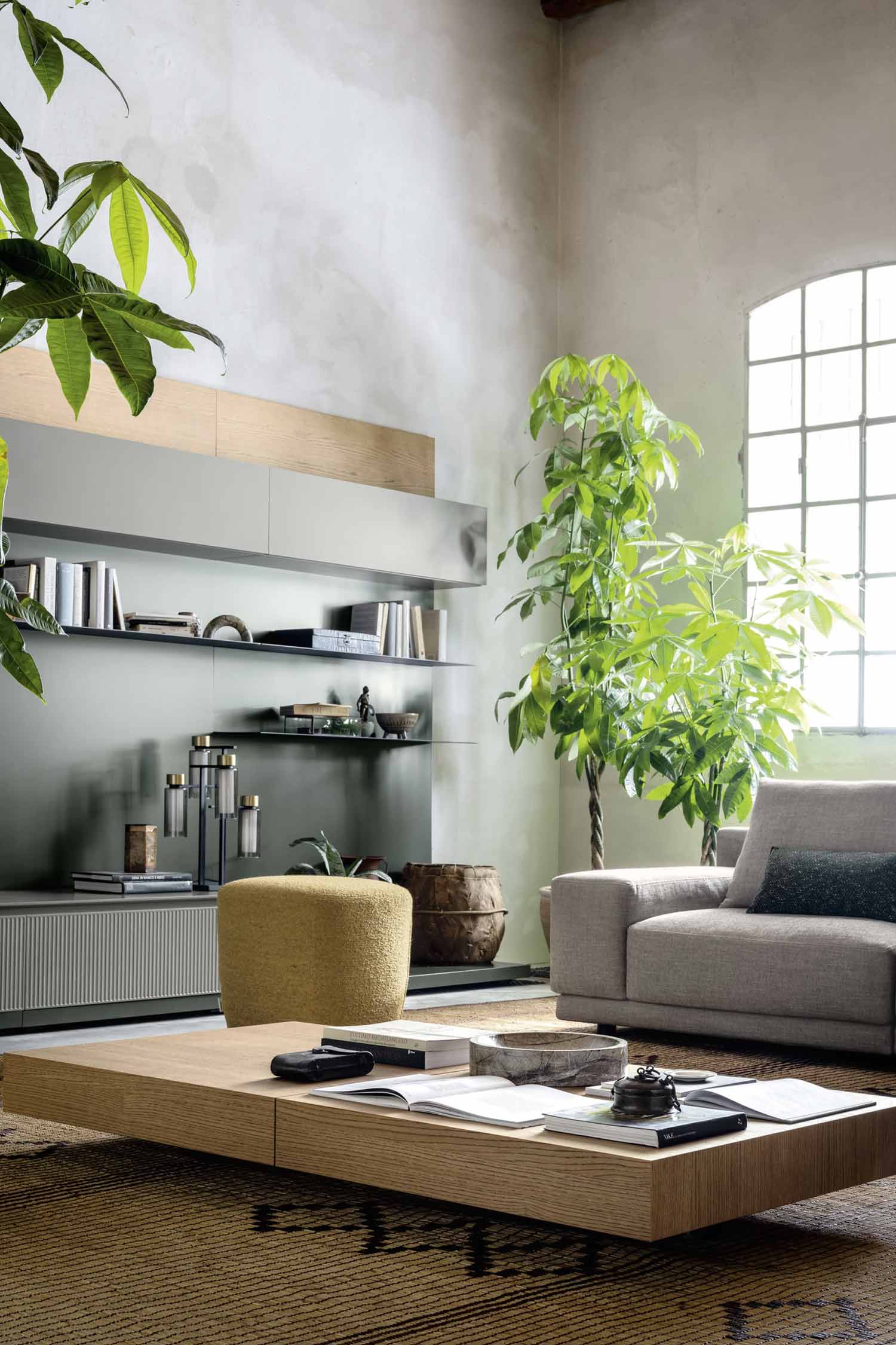 A cozy living room with a neutral-colored armchair, large potted plants, and a light wood coffee table with books and decorative objects on it. In the background, there's a gray wall-mounted unit with open shelving and a ribbed-front cabinet.
