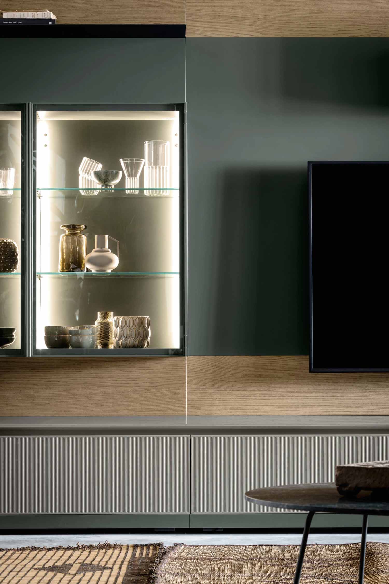A close-up of a dark green and light wood wall-mounted media unit. The unit features a glass-front display cabinet with internal lighting showcasing various glass and ceramic vessels. Below, a cabinet has a vertical ribbed texture. A television screen is visible on the right.