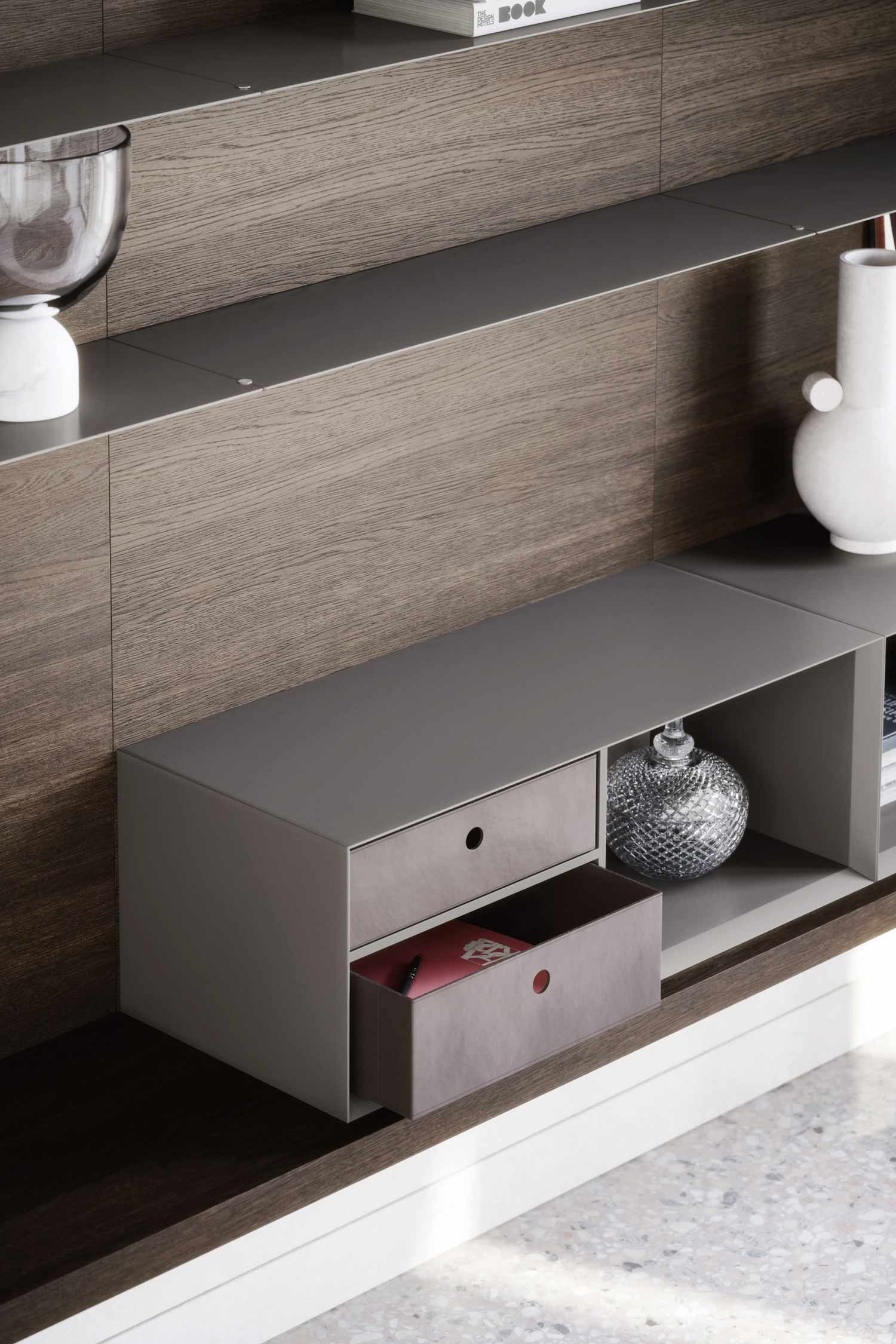 A close-up of a modern wall unit with a dark wood-paneled back. The unit features open gray shelves and a floating two-drawer cabinet. The bottom drawer is open, revealing a book, and a textured silver vase is on the shelf next to it.
