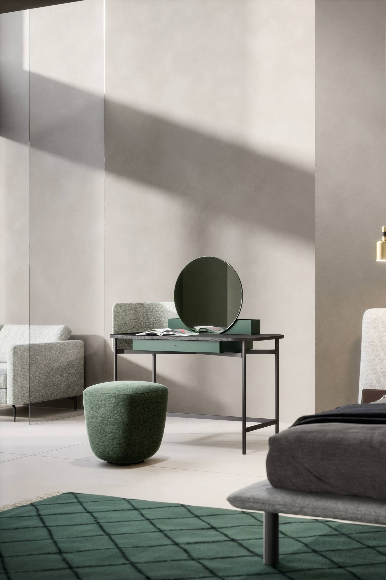 A contemporary bedroom or living space featuring a dark gray dressing table with a deep green circular mirror. An open book lies on the table, which also has a light gray upholstered privacy screen. In front of the table, a round, textured green ottoman provides seating. The room is decorated with a dark green patterned rug, and a light-colored sofa is partially visible in the background, bathed in sunlight.