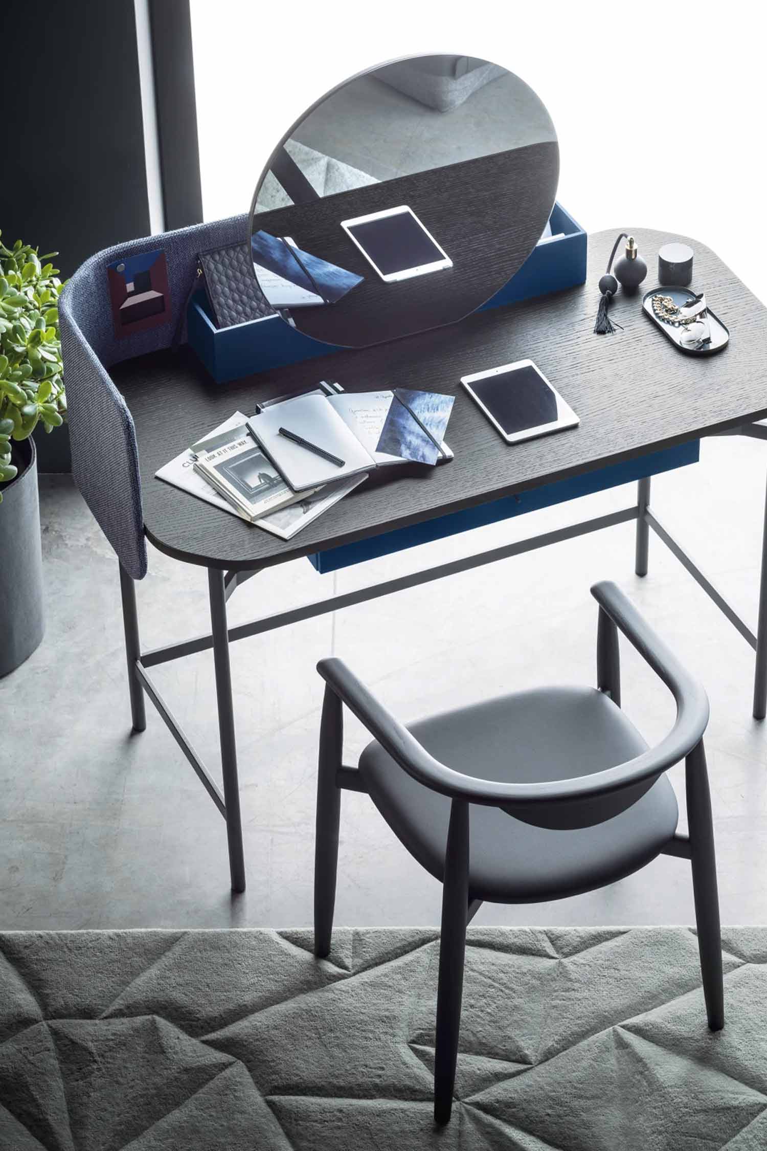 A stylish home office or vanity setup with a dark wooden desk and a curved, gray chair. The desk features a round mirror, an open notebook with a pen, a blue storage box containing various items, and a few small personal accessories. A potted plant sits to the left of the desk, and a textured gray rug is visible on the floor below.