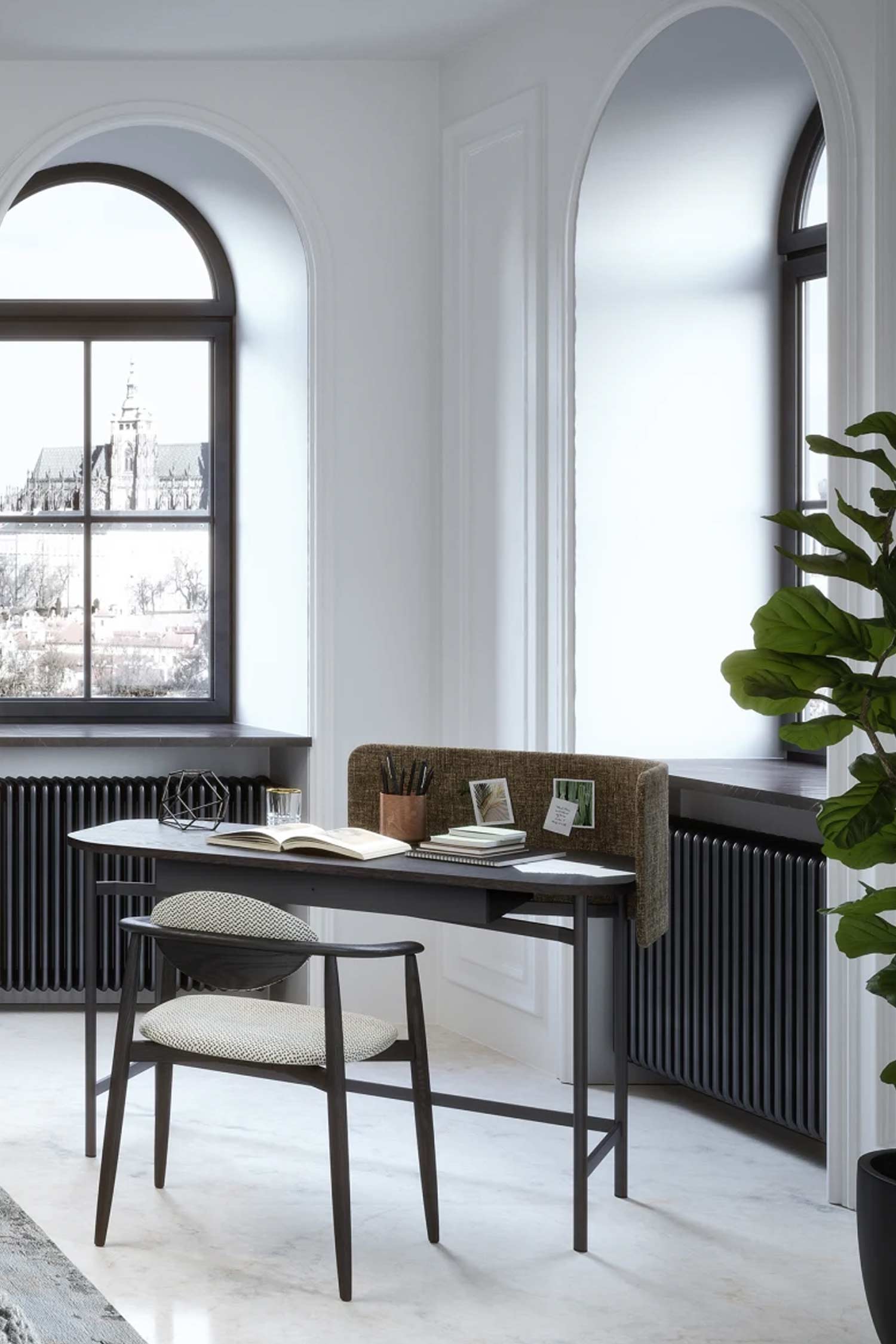 A minimalist home office with a dark wood desk and a light gray upholstered privacy screen. An open notebook, a pen holder, and a glass of water are arranged on the desk. A stylish dark wooden chair with a white speckled cushion is tucked underneath. The room is brightly lit and features two large arched windows with black radiators below them. One window looks out onto a cityscape. A large potted plant is visible in the foreground on the right.