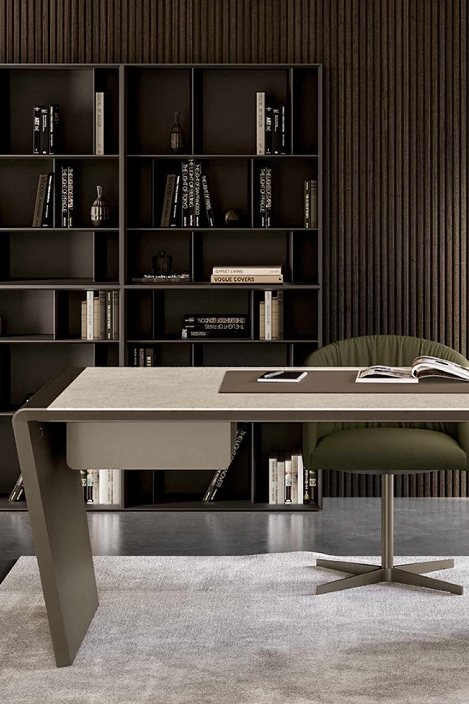 A designer office desk with a light top and dark taupe leg, seen from the front. The desk is furnished with a leather pad, a magazine, and a phone. A dark green office chair is partially visible against a background of a large, dark wood modular bookshelf and a slatted accent wall.