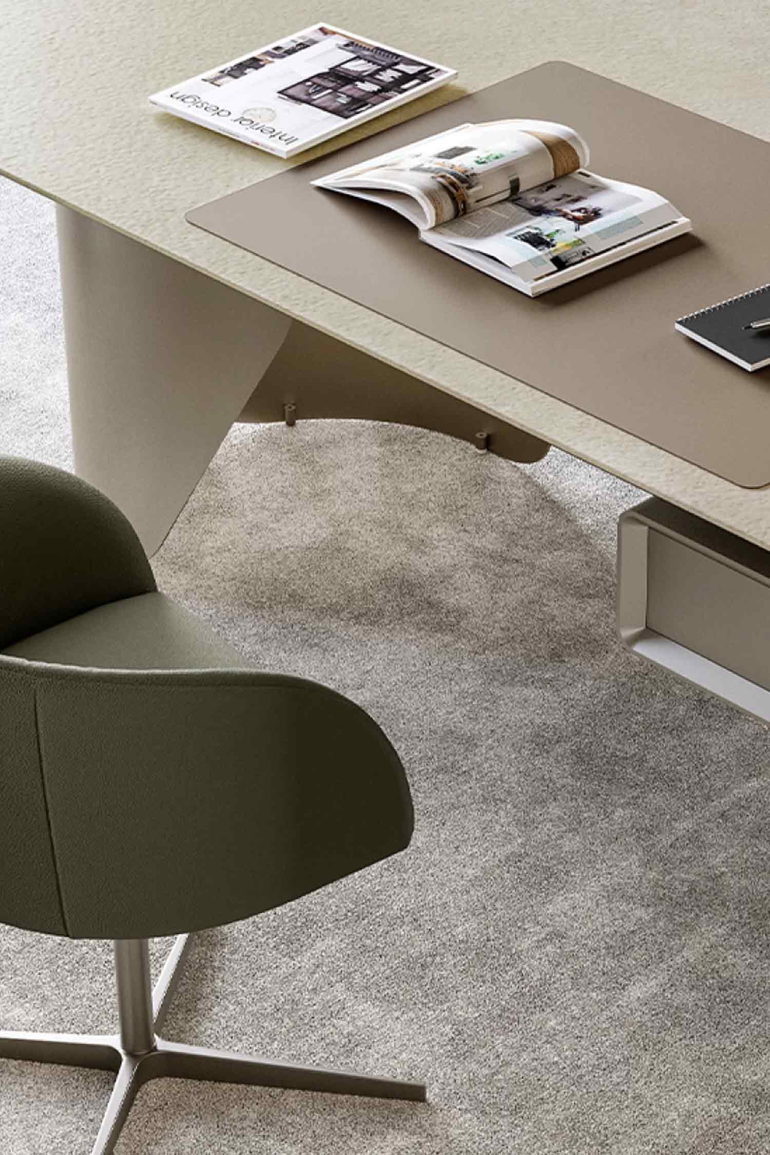 A high-angle, close-up view of a modern office desk with a light beige, textured top and a taupe leather desk pad. An open "Interior Design" magazine and a notebook are on the desk. A dark olive green swivel chair with a metal base is in the foreground, all set on a gray carpet.