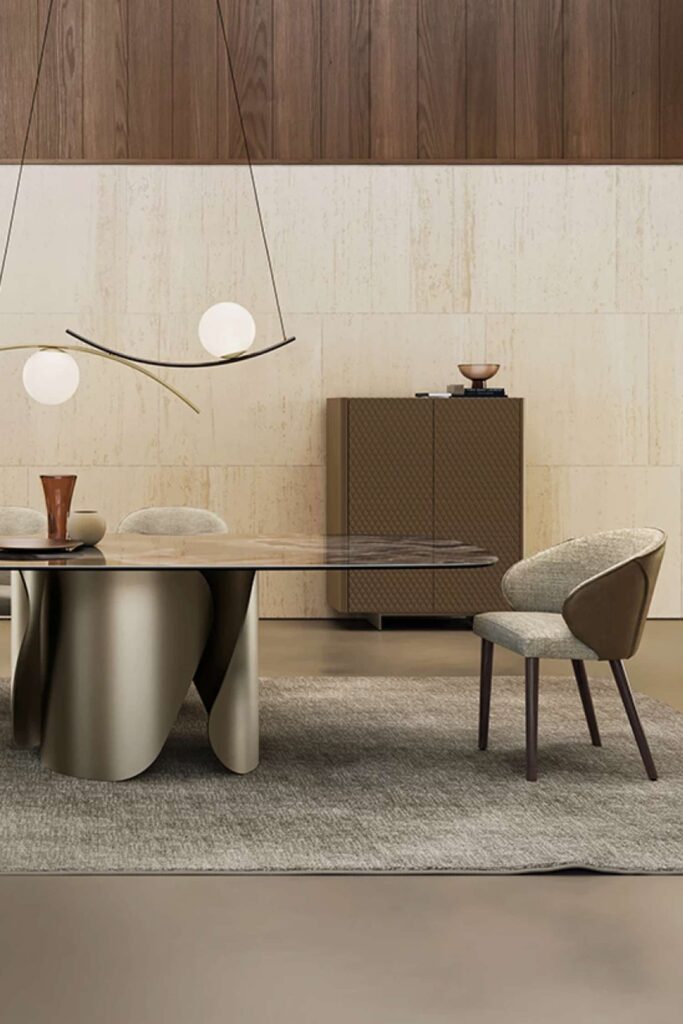 Full view of the Onda dining table with a glossy stone top and bronze-toned metallic base, surrounded by upholstered dining chairs, an overhead modern curved pendant light, and a patterned cabinet in a spacious, light-colored room.