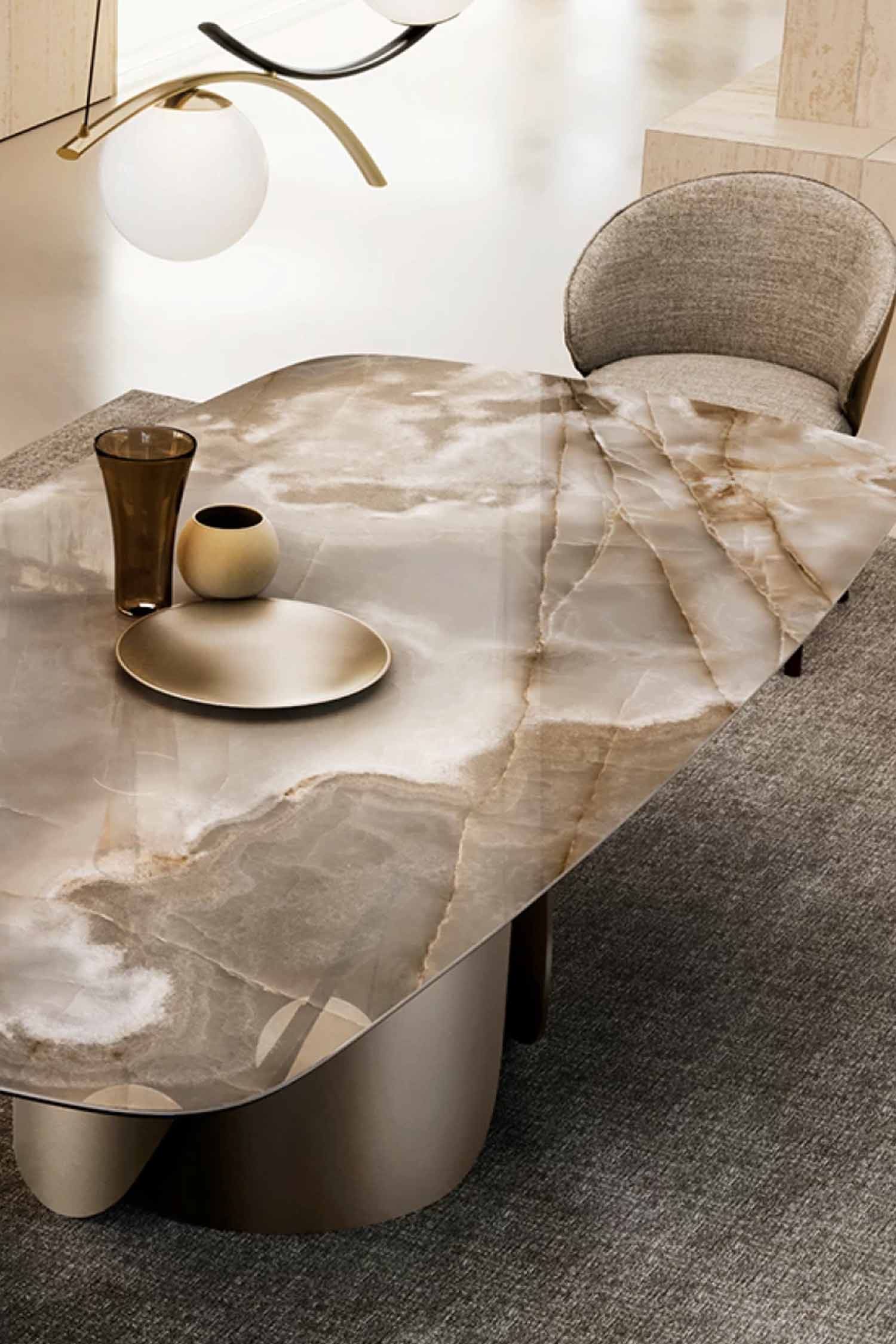 Close-up, angled view of the Onda dining table's glossy top made of richly veined natural onyx or marble in warm taupe and white tones, with a decorative gold plate and vase, and a gray upholstered chair visible.