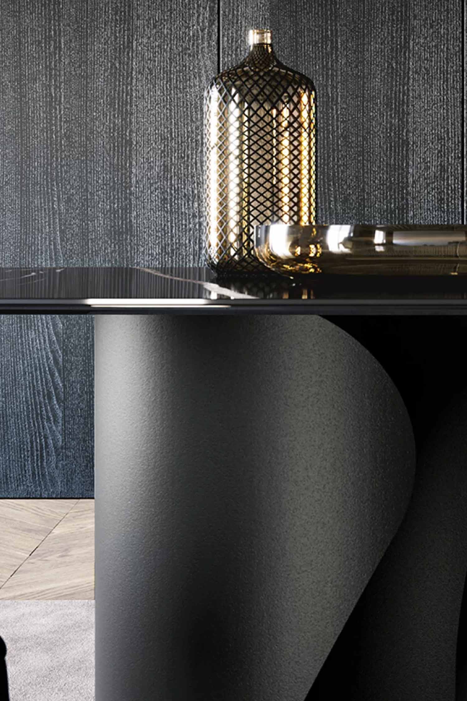 Vertical close-up detail shot of the Onda table, highlighting the contrast between the glossy black tabletop, the matte sculptural black base, and a tall, geometric gold vase, set against a dark paneled wall.
