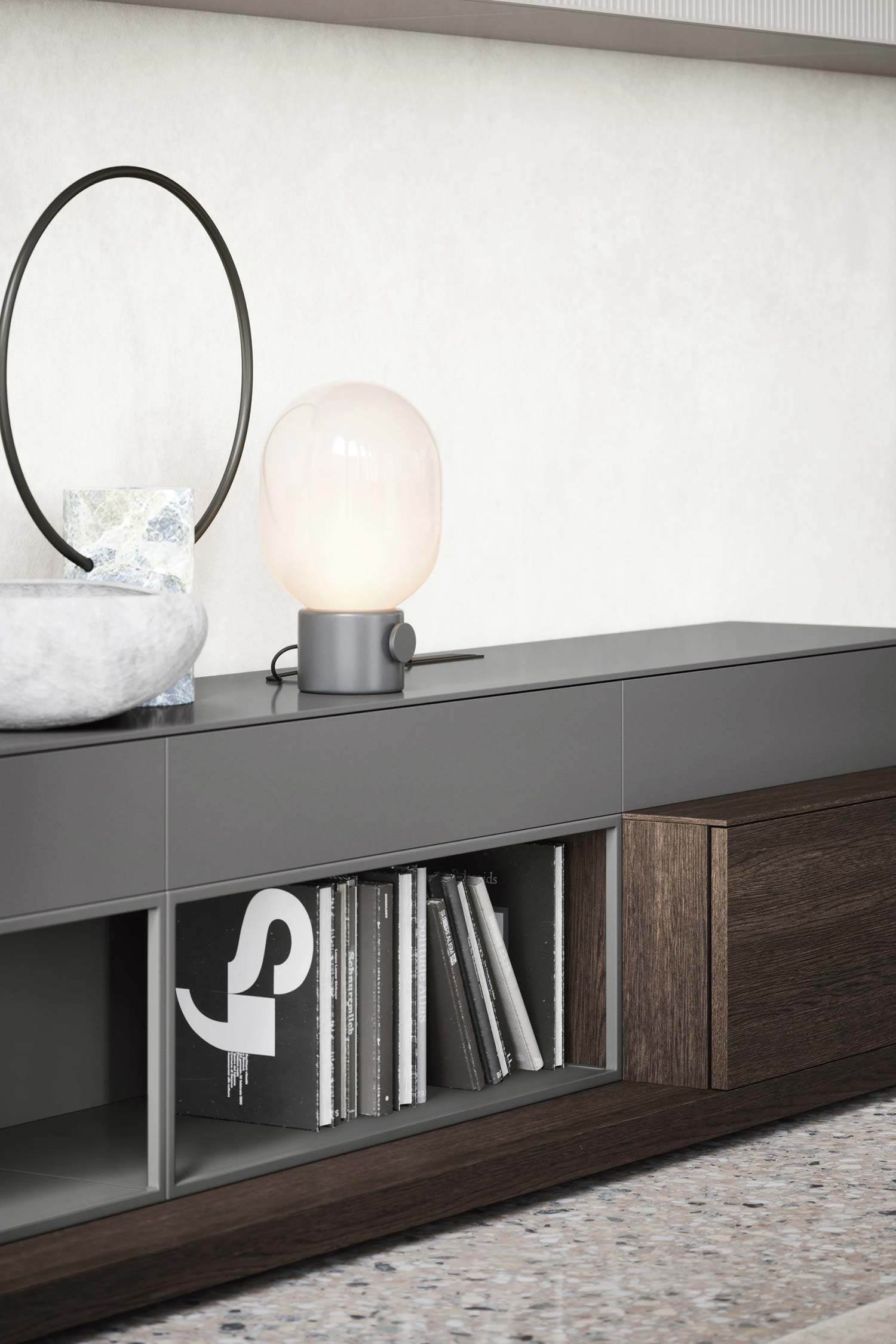 A close-up, low-angle shot of a modern gray credenza with a dark wooden drawer. A section of the open shelving is visible, showing a stack of books. A contemporary white globe lamp sits on top of the credenza.
