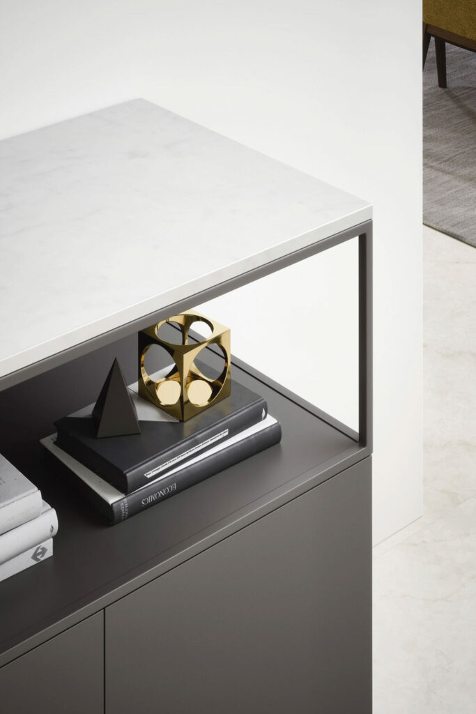 A close-up of a modern, two-toned credenza. The top shelf has a white marble surface, and a smaller, gray open shelf below holds books and a gold cube-shaped decorative sculpture.