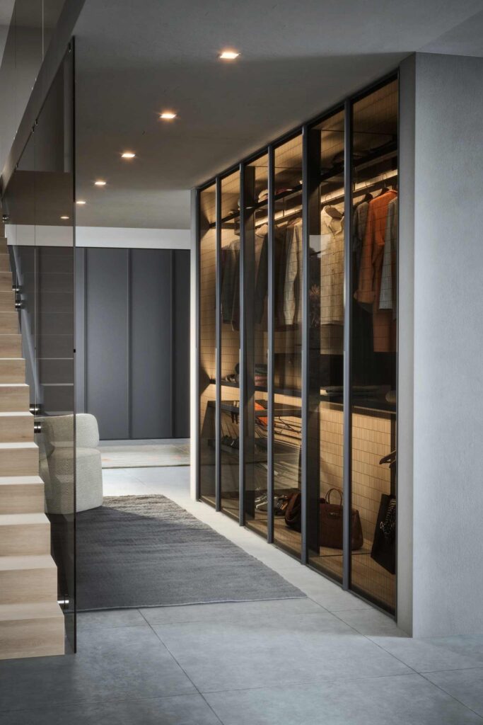 A modern walk-in closet or hallway with a large, hinged wardrobe featuring a dark frame and a translucent, ribbed glass front. The wardrobe is internally lit, showing a mix of hanging clothes, folded items, and a bag on a shelf. A light grey rug is on the concrete floor, leading toward a plush, light-colored chair. In the background, a staircase with wooden steps and a glass railing is visible on the left, and a dark wall with a flat-panel wardrobe is on the right.