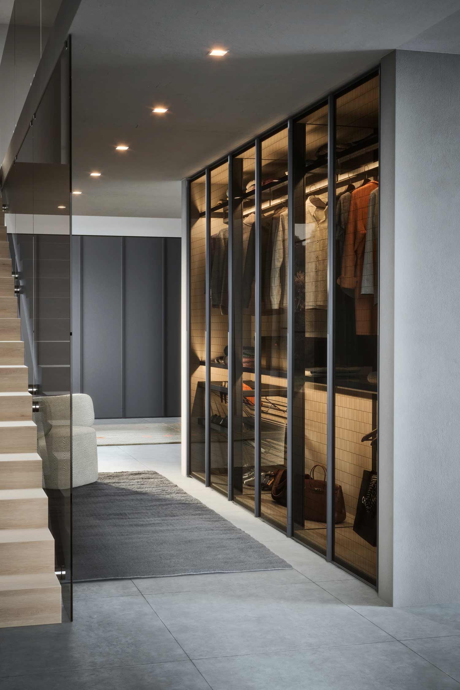 A modern walk-in closet or hallway with a large, hinged wardrobe featuring a dark frame and a translucent, ribbed glass front. The wardrobe is internally lit, showing a mix of hanging clothes, folded items, and a bag on a shelf. A light grey rug is on the concrete floor, leading toward a plush, light-colored chair. In the background, a staircase with wooden steps and a glass railing is visible on the left, and a dark wall with a flat-panel wardrobe is on the right.