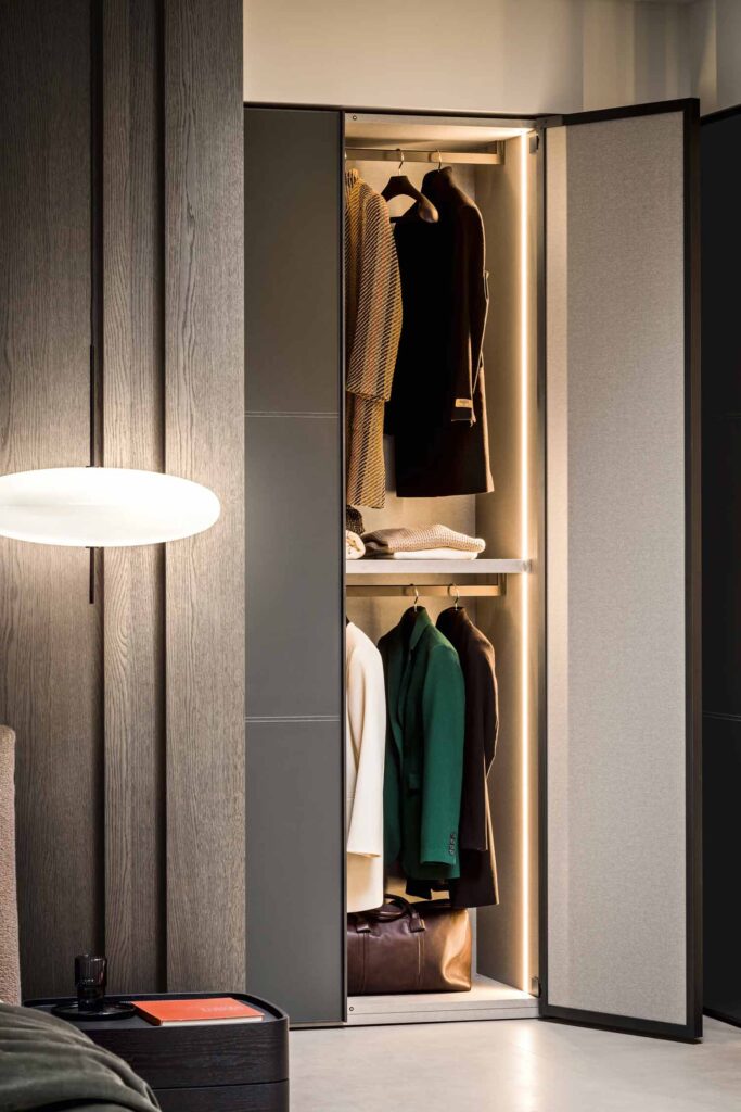 A close-up view of a luxury wardrobe with two open hinged doors. The door on the right has a light grey fabric finish, and the door on the left has a dark grey finish. The interior is brightly lit with LED strips, showcasing hanging clothes on two levels. A brown leather bag rests on the floor inside the wardrobe. The wardrobe is set against a textured, dark wood panel wall. A modern, oval-shaped wall light is mounted on the wall to the left, and a dark side table with a book on it is visible in the foreground.