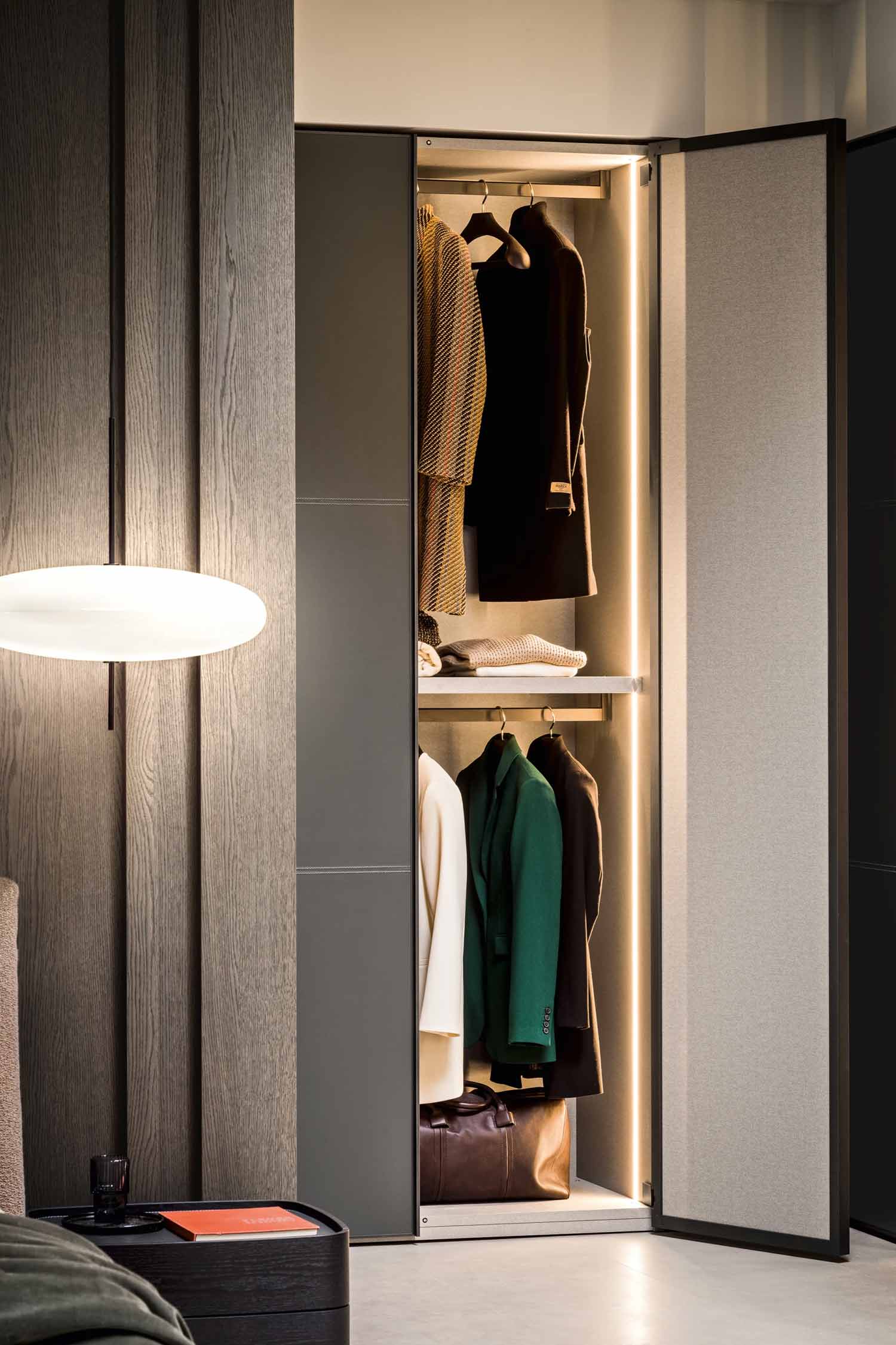 A close-up view of a luxury wardrobe with two open hinged doors. The door on the right has a light grey fabric finish, and the door on the left has a dark grey finish. The interior is brightly lit with LED strips, showcasing hanging clothes on two levels. A brown leather bag rests on the floor inside the wardrobe. The wardrobe is set against a textured, dark wood panel wall. A modern, oval-shaped wall light is mounted on the wall to the left, and a dark side table with a book on it is visible in the foreground.