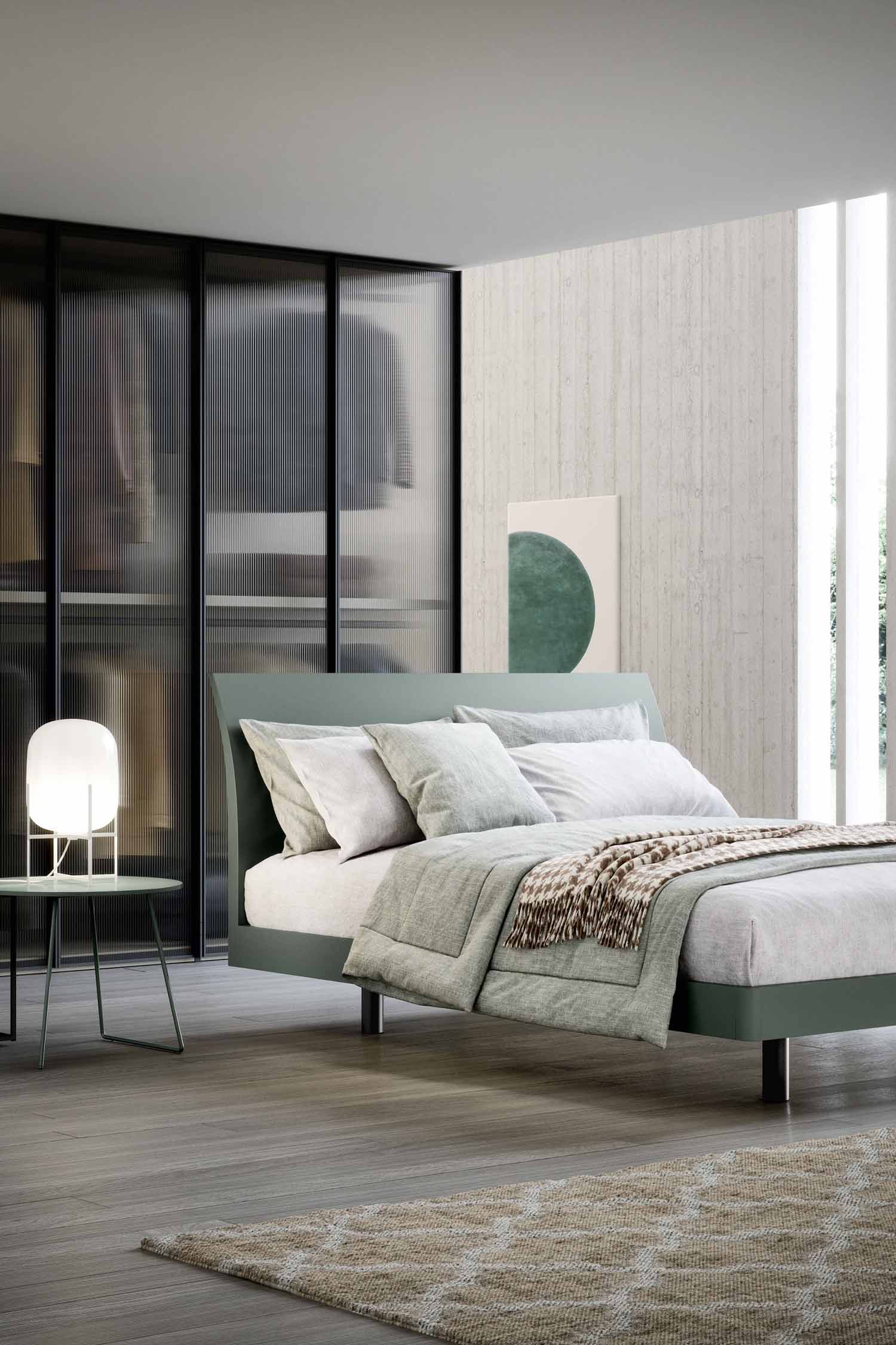 A minimalist bedroom featuring a low-profile green upholstered bed with grey and white bedding. A small side table with a modern white glowing lamp sits to the left. The room has a dark wood floor with a textured, light-colored rug. In the background, there's a large, wall-to-wall wardrobe with a dark frame and translucent, ribbed glass sliding doors. A piece of art with a green circular pattern hangs on a bare concrete wall to the right. The room is filled with natural light from a large window.