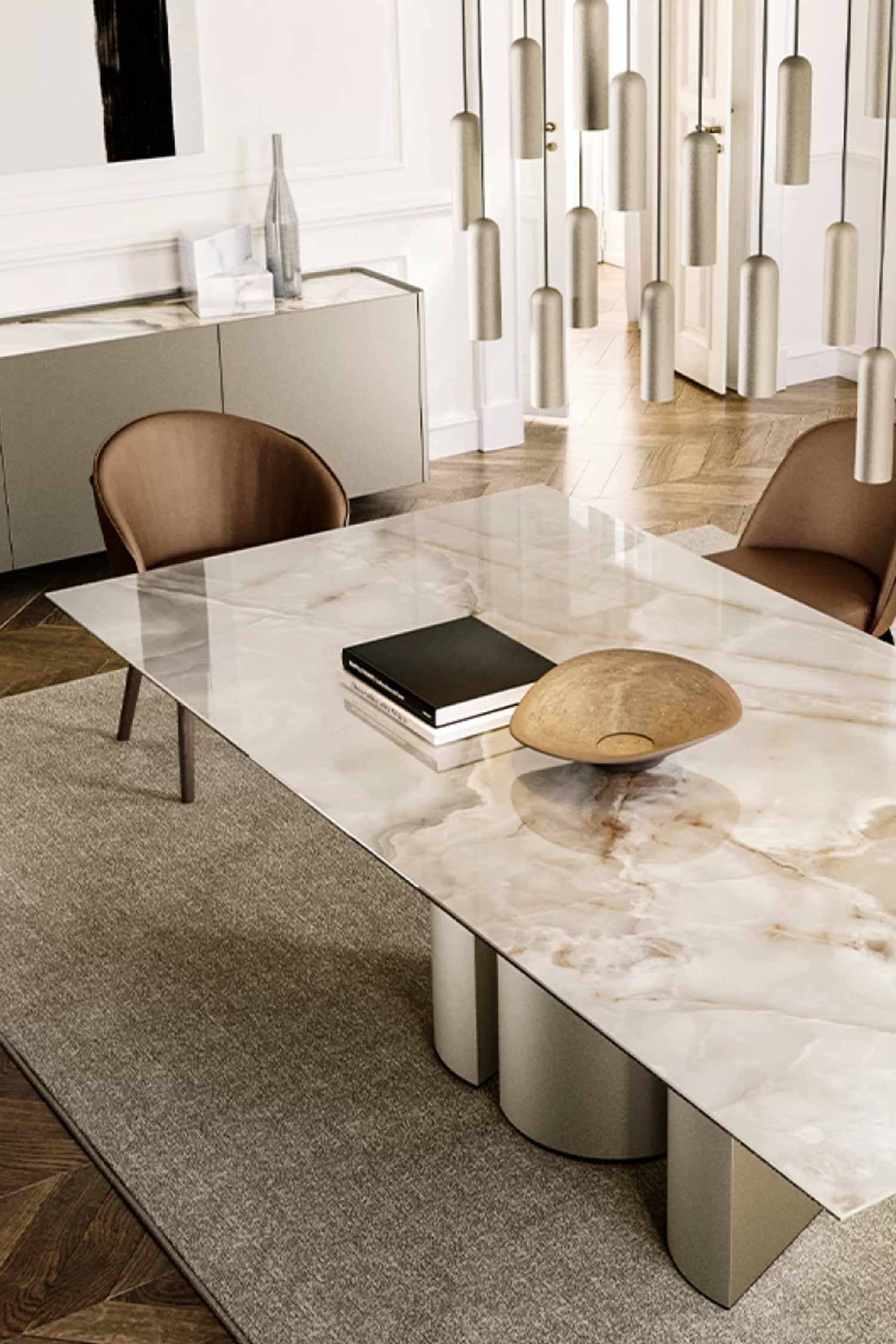 Close-up of a glossy white marble dining tabletop with subtle tan veining, featuring a decorative wooden bowl and books, showing the taupe cylindrical base of the table, and brown leather chairs with a matching sideboard in the background.