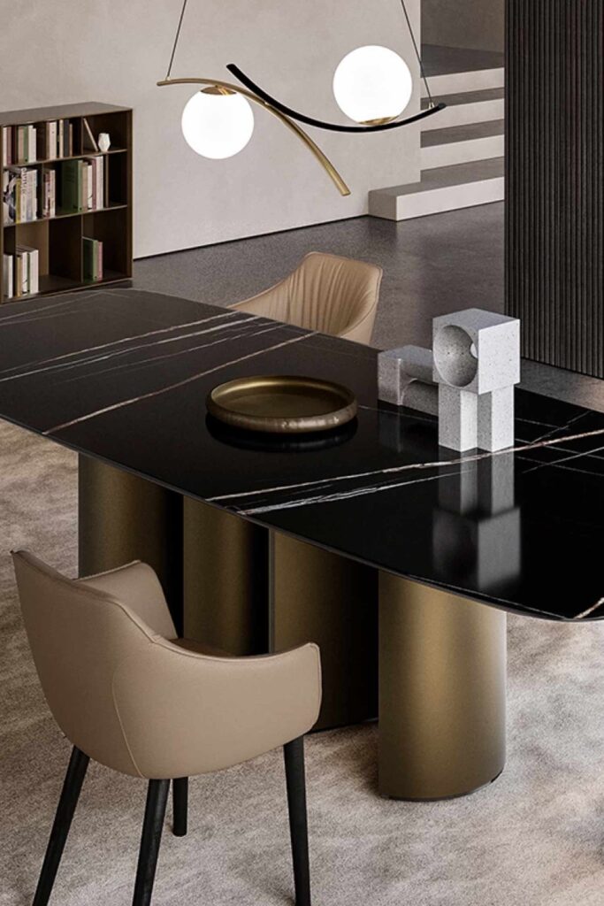 Close-up of a black marble dining table with white veining, highlighting the metallic gold pedestal legs and decorative objects (a gold tray and geometric stone sculptures) on the table, with light beige chairs and a modern light fixture visible.
