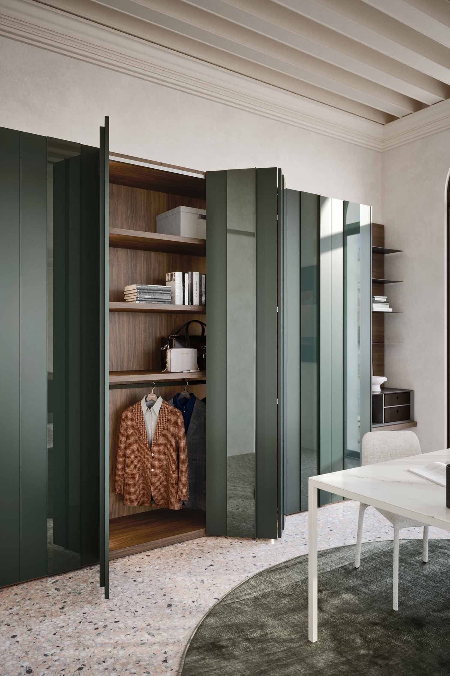 A modern office space featuring a sage green hinged wardrobe that is partially open. The wardrobe's interior is dark wood with shelves holding books and a white purse. A blazer hangs from a rod below the shelves. The wardrobe doors have mirrored strips that reflect the room. A white desk and chair are visible on the right, along with a dark wood and metal bookshelf against the wall. The room has a light terrazzo floor and a dark, circular rug.