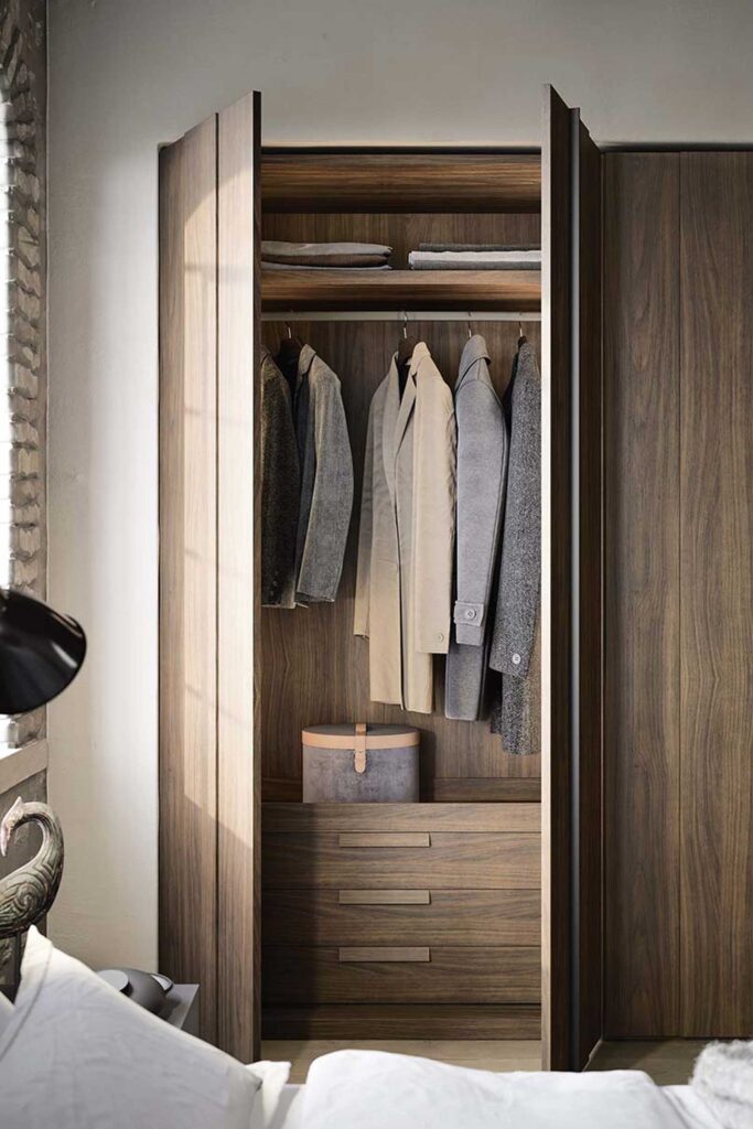 A close-up of a modern, open wardrobe with a dark wood interior. The wardrobe has a full-length section for hanging clothes, with jackets and coats visible. Below the hanging section are three drawers and a gray, rectangular box with a brown leather strap handle