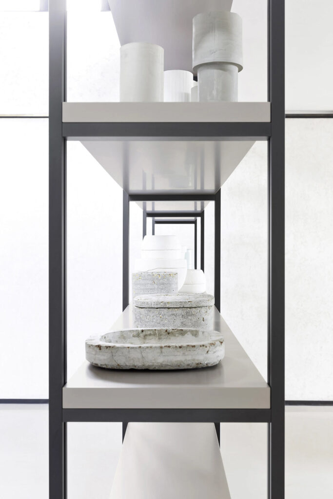 A close-up, eye-level shot of a modern shelving unit with dark grey frames and light grey shelves. The shelves are sparsely decorated with various white and grey ceramic or stone decorative objects, including cylindrical forms and textured dishes, all displayed against a bright, white background. The perspective gives the impression of a long, receding set of shelves.