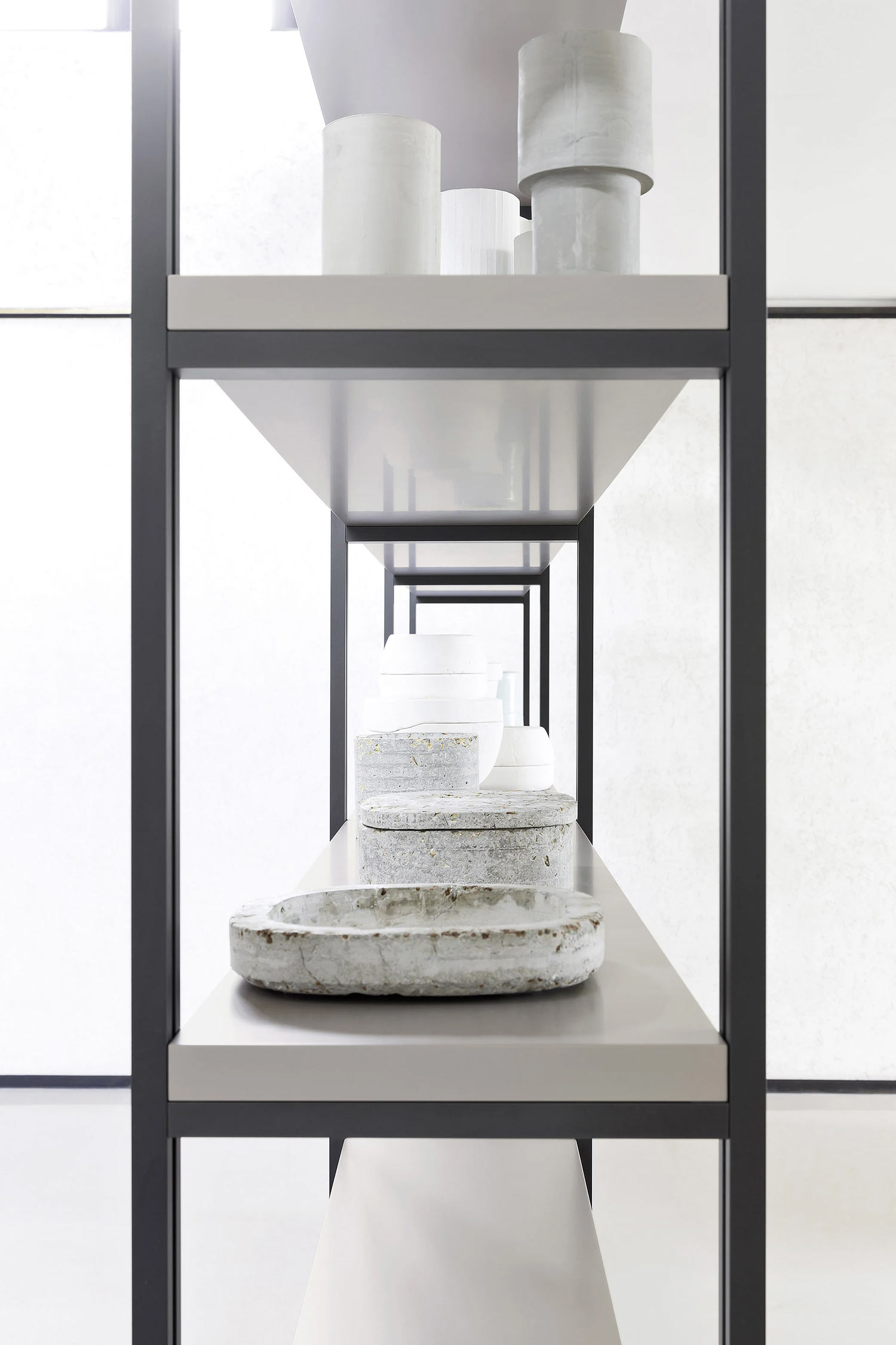 A close-up, eye-level shot of a modern shelving unit with dark grey frames and light grey shelves. The shelves are sparsely decorated with various white and grey ceramic or stone decorative objects, including cylindrical forms and textured dishes, all displayed against a bright, white background. The perspective gives the impression of a long, receding set of shelves.