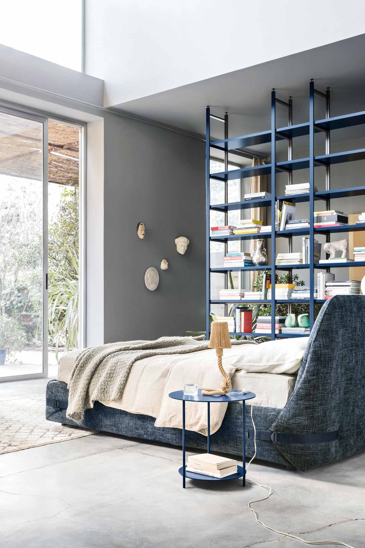 A contemporary bedroom with a large bed in the foreground. The bed is upholstered in a textured blue fabric and has a light beige throw blanket. A round, blue side table with two tiers sits next to the bed, holding a small lamp and a glass of water. In the background, a blue open-shelving bookcase acts as a room divider, separating the sleeping area from the rest of the room. The room has grey walls, a concrete floor, and a large sliding glass door that leads to an outdoor patio.
