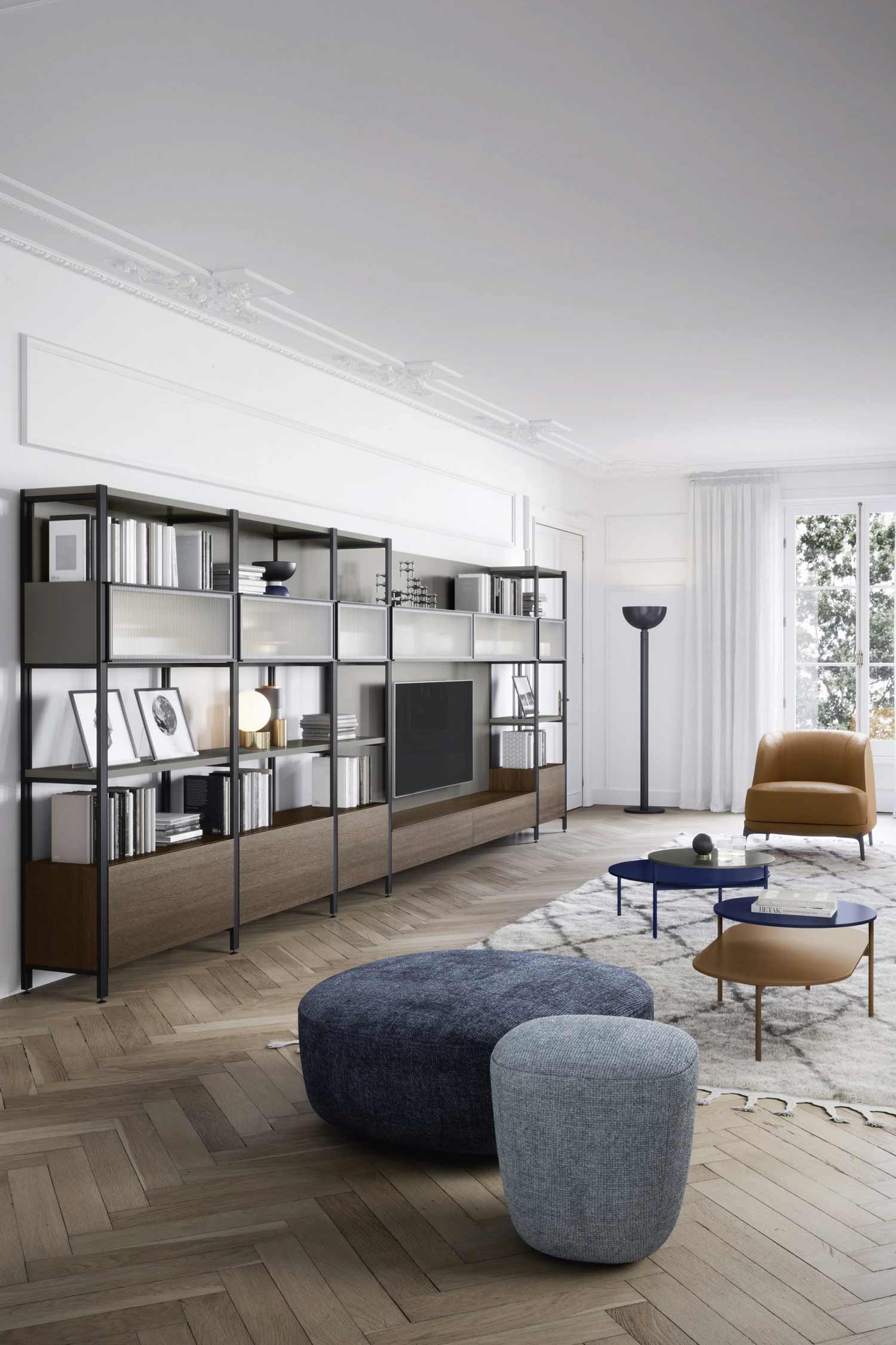 A stylish living room featuring a long, black metal and wood media unit with integrated shelving. The unit spans the wall, displaying a television in the center, surrounded by books and decorative items on shelves with clear and fluted glass fronts. In the foreground, there are two round, upholstered poufs in blue and grey. A pair of oval-shaped, two-tiered coffee tables in blue and tan sit on a textured white rug. The room has light wood floors, high ceilings, a brown leather armchair, and a large window with white curtains