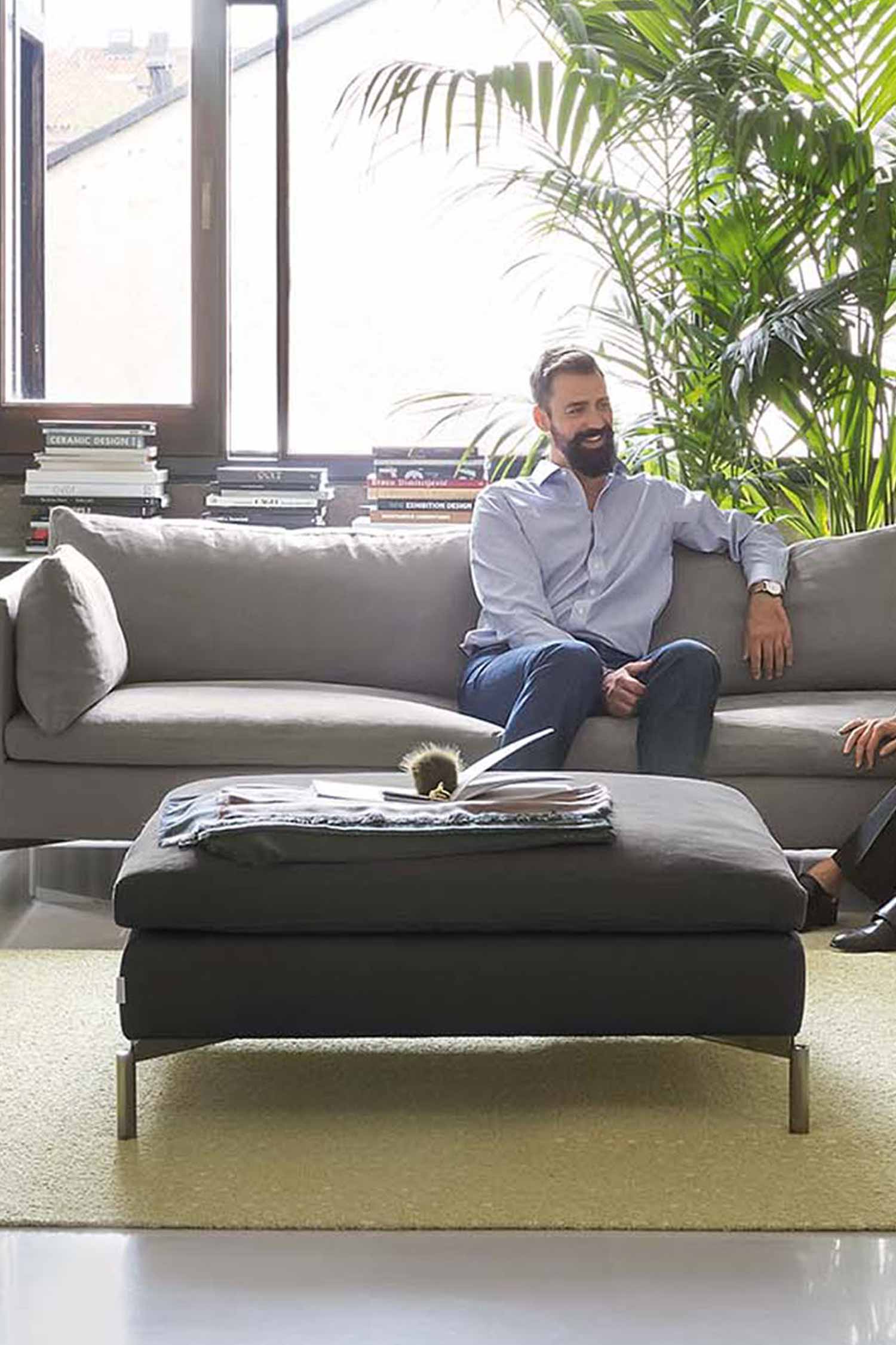 A low-angle, eye-level shot of a man relaxing on a large, light gray sofa in a modern living room. He is sitting on the left side of the sofa with his arm on the backrest, smiling. In the foreground, a large, dark gray ottoman with thin metal legs is centered on a light green rug. A few magazines and a small decorative object are on top of the ottoman. The background features a large window with lush green plants behind it. A stack of books is on a side table on the far left.
