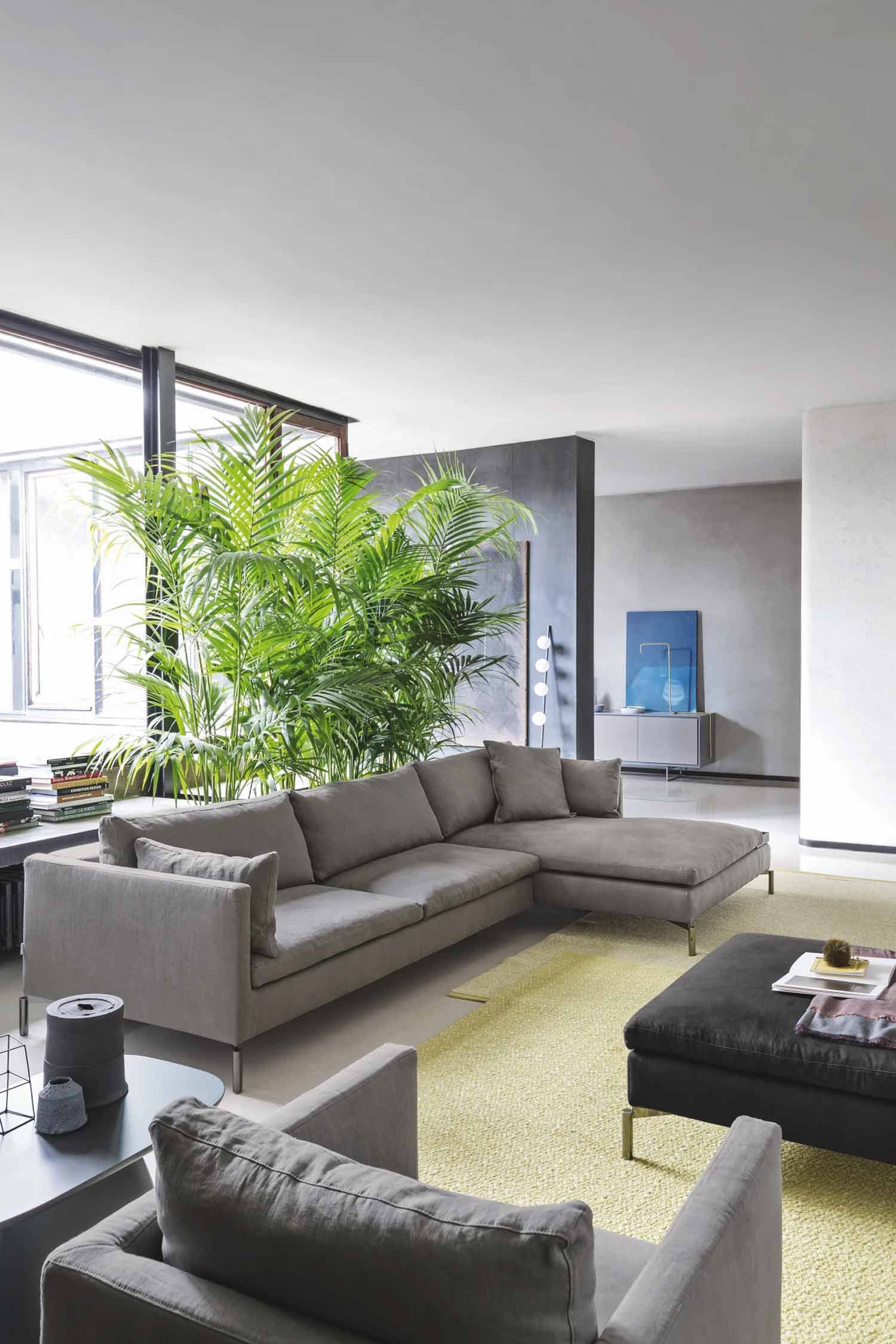 A low-angle, wide shot of a spacious living room. A large, dark blue upholstered armchair with a unique rounded shape is in the foreground on the left, next to a multi-tiered black coffee table. On the right, a light gray ottoman is partially visible, with a dark blue blanket draped over it. In the center, a tall floor lamp with a gold stem and three circular white lights stands between the chair and the ottoman. The room has light gray walls, dark wood flooring, and large white double doors in the background.