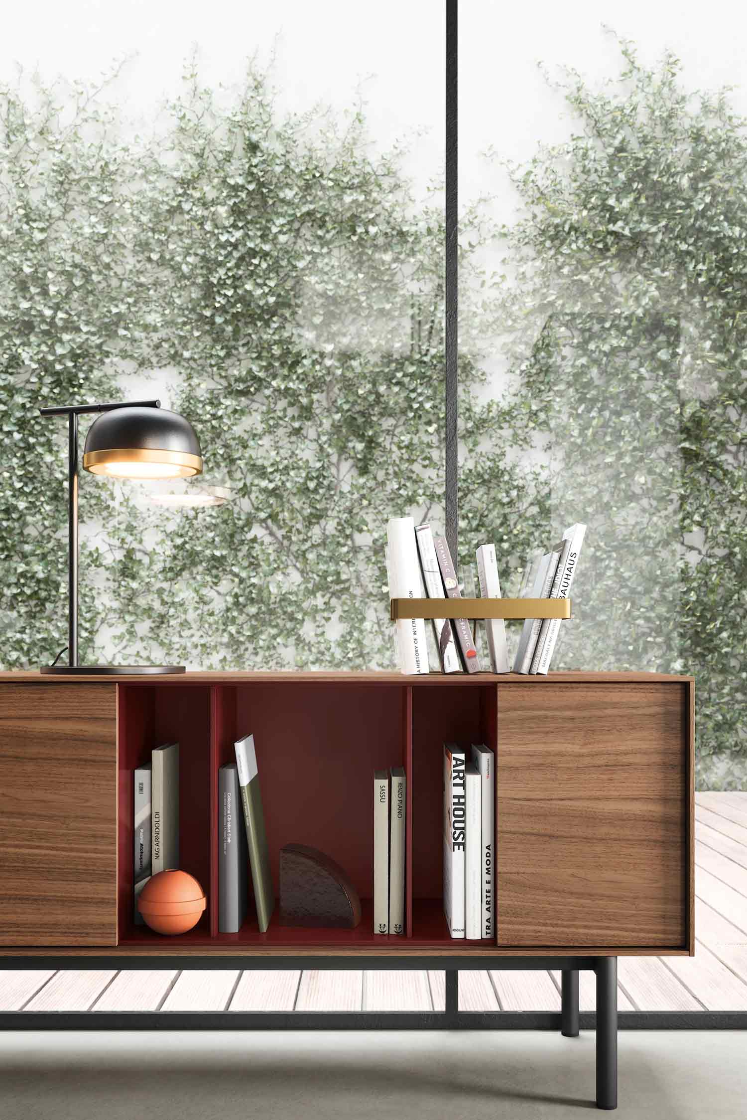 A detail shot of a modern wooden sideboard with a rich red interior and a brass-colored metal handle. The sideboard has two wood veneer sliding doors and two open compartments. The compartments are filled with books and a small orange ball. A stylish black and gold lamp is on top of the sideboard, and in the background, a large window reveals a wall covered in green ivy.