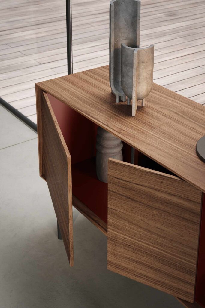 A close-up of a modern wooden sideboard with two open doors, revealing a dark red interior. The sideboard has a rich wood grain finish and stands on dark metal legs. Various decorative vases and objects are visible both inside and on top of the cabinet. The background shows a light-colored wooden patio deck and a floor-to-ceiling window.
