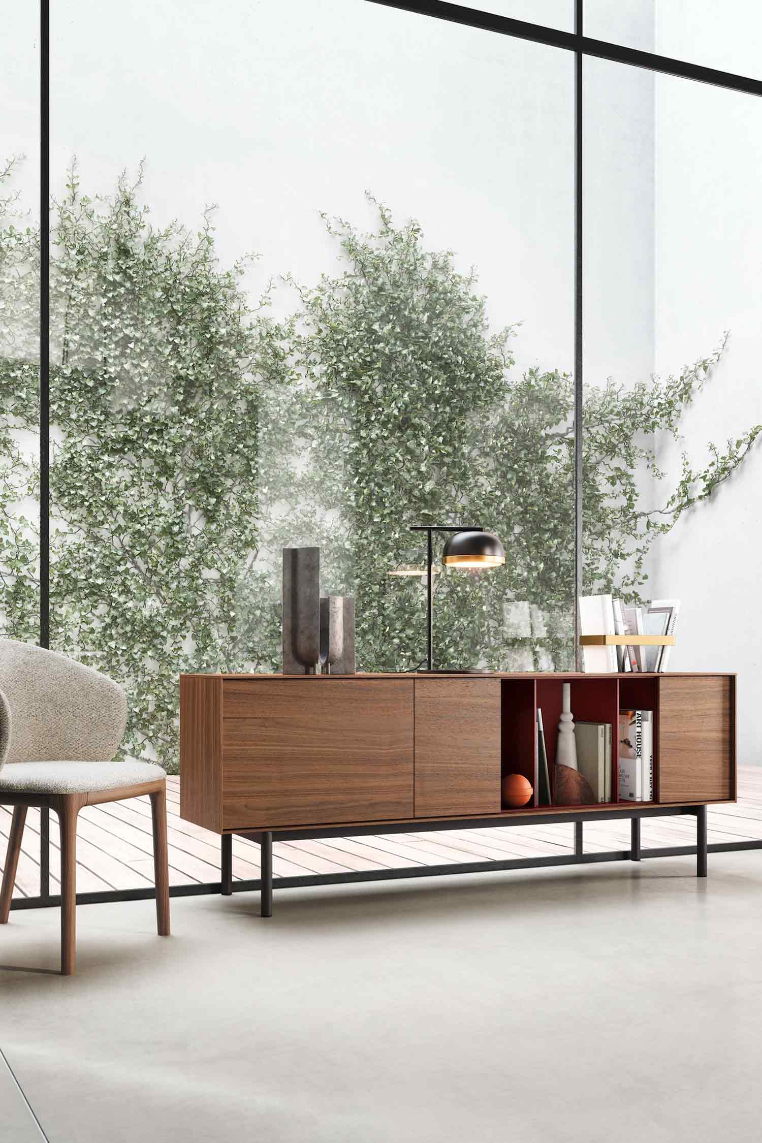 A stylish, modern living space featuring a long wooden sideboard with a dark red interior, two closed cabinets, and two open shelves filled with books and a decorative orange ball. On top of the sideboard are two dark grey vases and a black and gold table lamp. To the left, a light-colored upholstered chair with wooden legs is partially visible. Large floor-to-ceiling windows behind the sideboard look out onto an outdoor area with a wall covered in green ivy and a light-colored wooden deck. The interior floor is a light grey.
