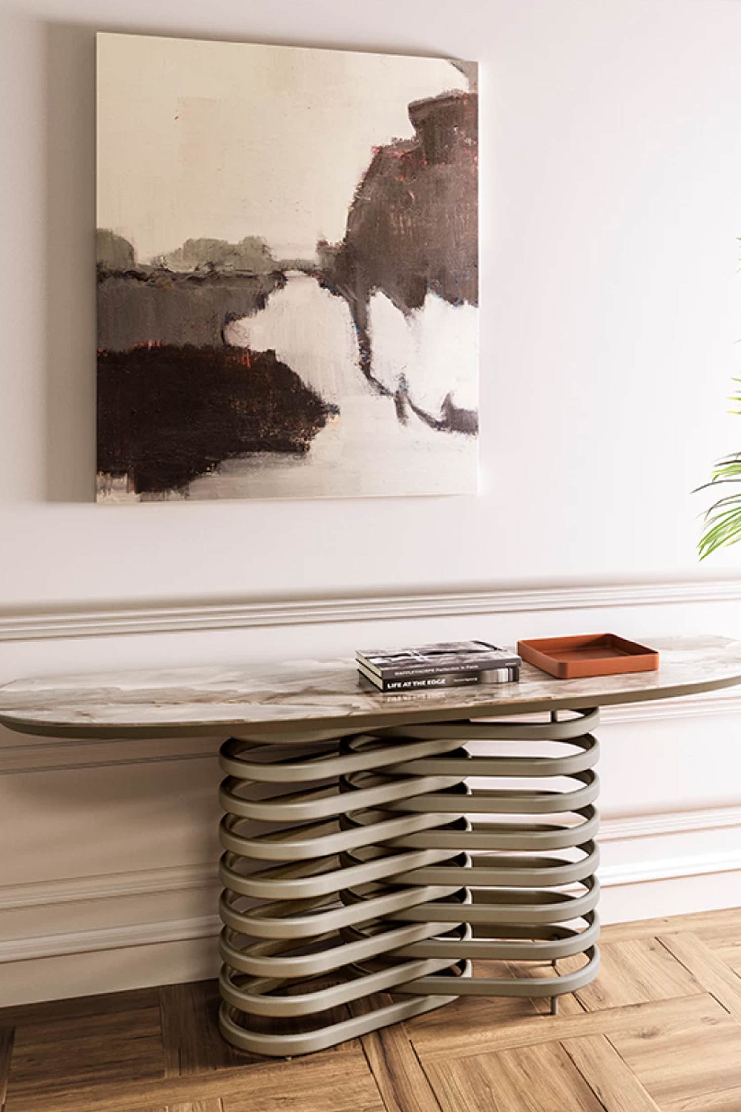 Full-length view of the Rotolo console table, featuring an oblong, veined marble top and a coiled, interwoven tan metal base, positioned against a white wall with wainscoting. A dark-toned abstract painting hangs above the table, which holds a book and a small tray.