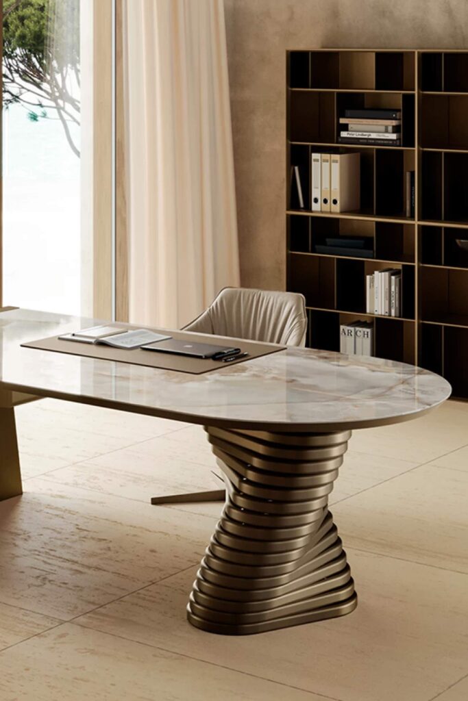 Detailed view of the Rotolo desk's marble top and its twisted metal base. A leather desk pad, papers, and a laptop are on the desk, with a geometric, open-shelf bookcase visible in the background.