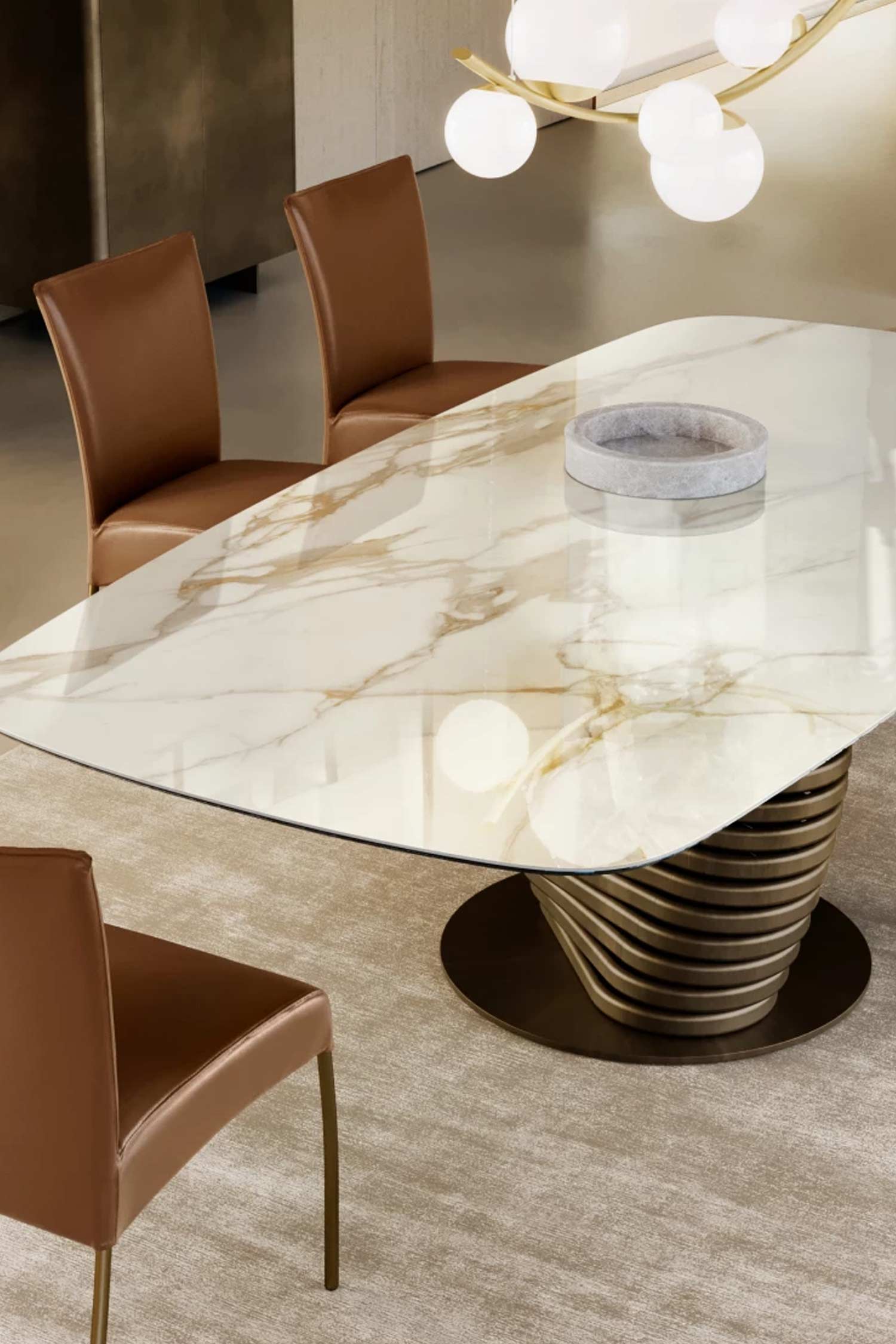 Close-up detail of the oval Rotolo dining table's glossy white top with prominent golden-brown marble veining. The unique bronze twisted base is clearly visible, surrounded by tan leather dining chairs on a light-colored rug.