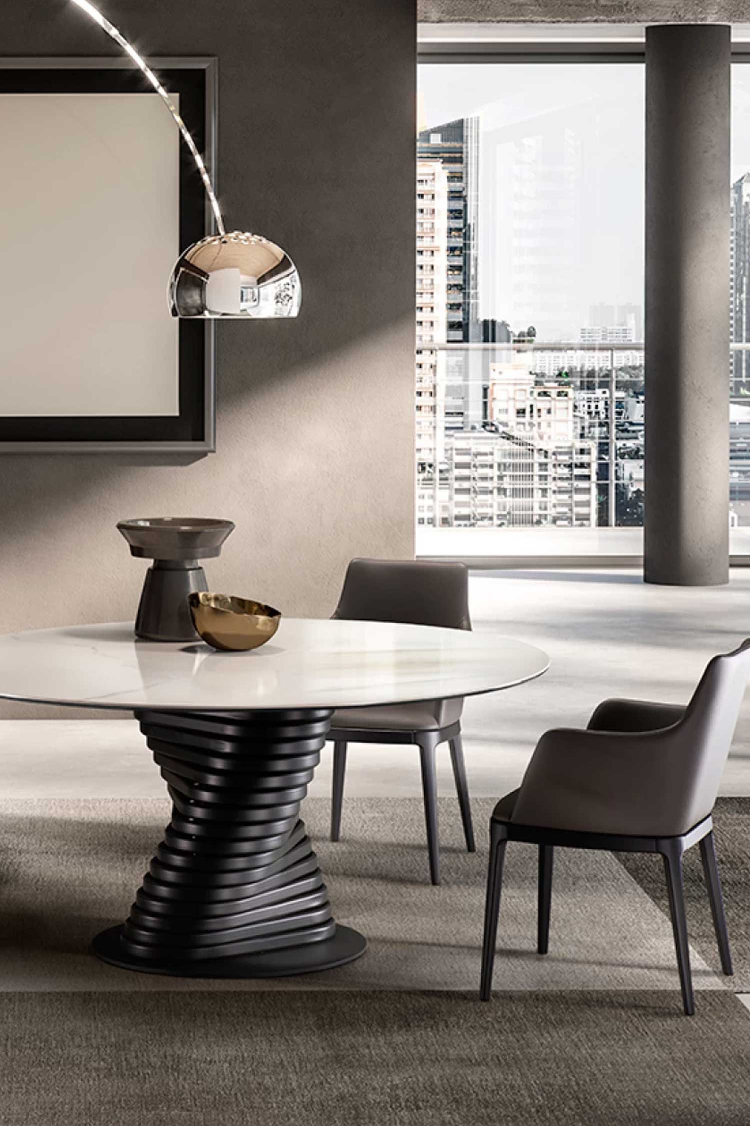 Detail shot of a round Rotolo dining table with a white marble top and a black twisted base. Two dark gray leather chairs are visible, with a modern pendant light and a window overlooking a city skyline in the background.