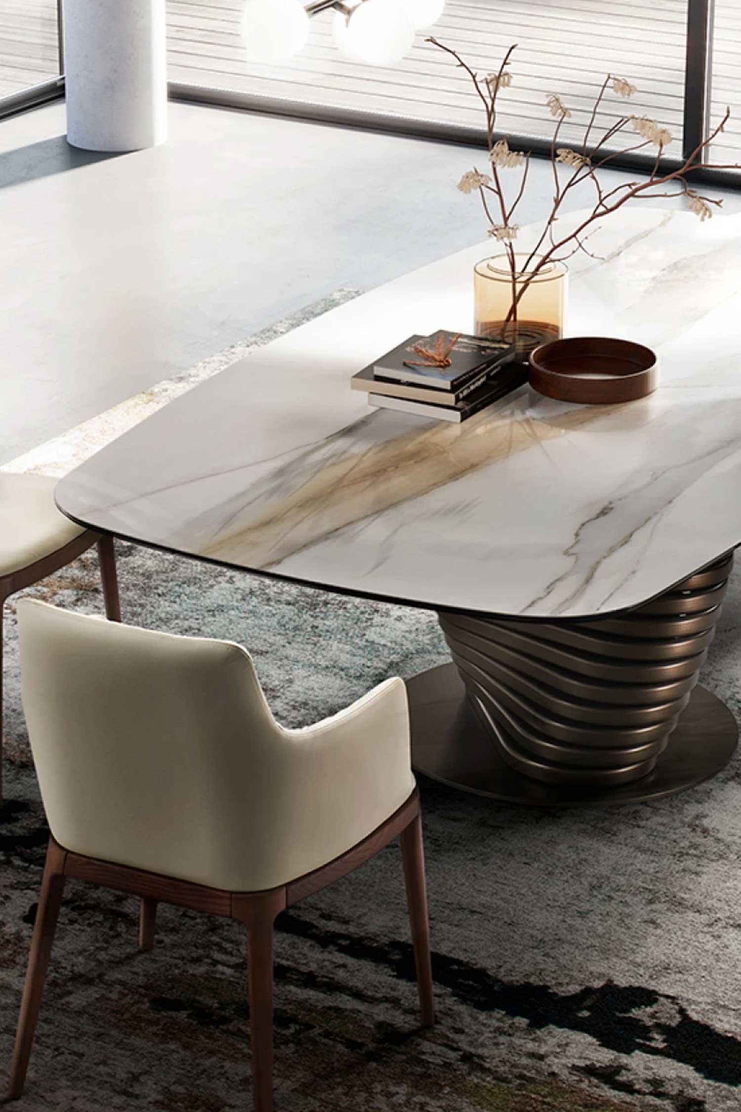Overhead detail view of the oval Rotolo dining table with a white marble top and a dark bronze twisted base. The table is styled with books and a vase, and a cream-colored dining chair is in the foreground on a patterned rug.