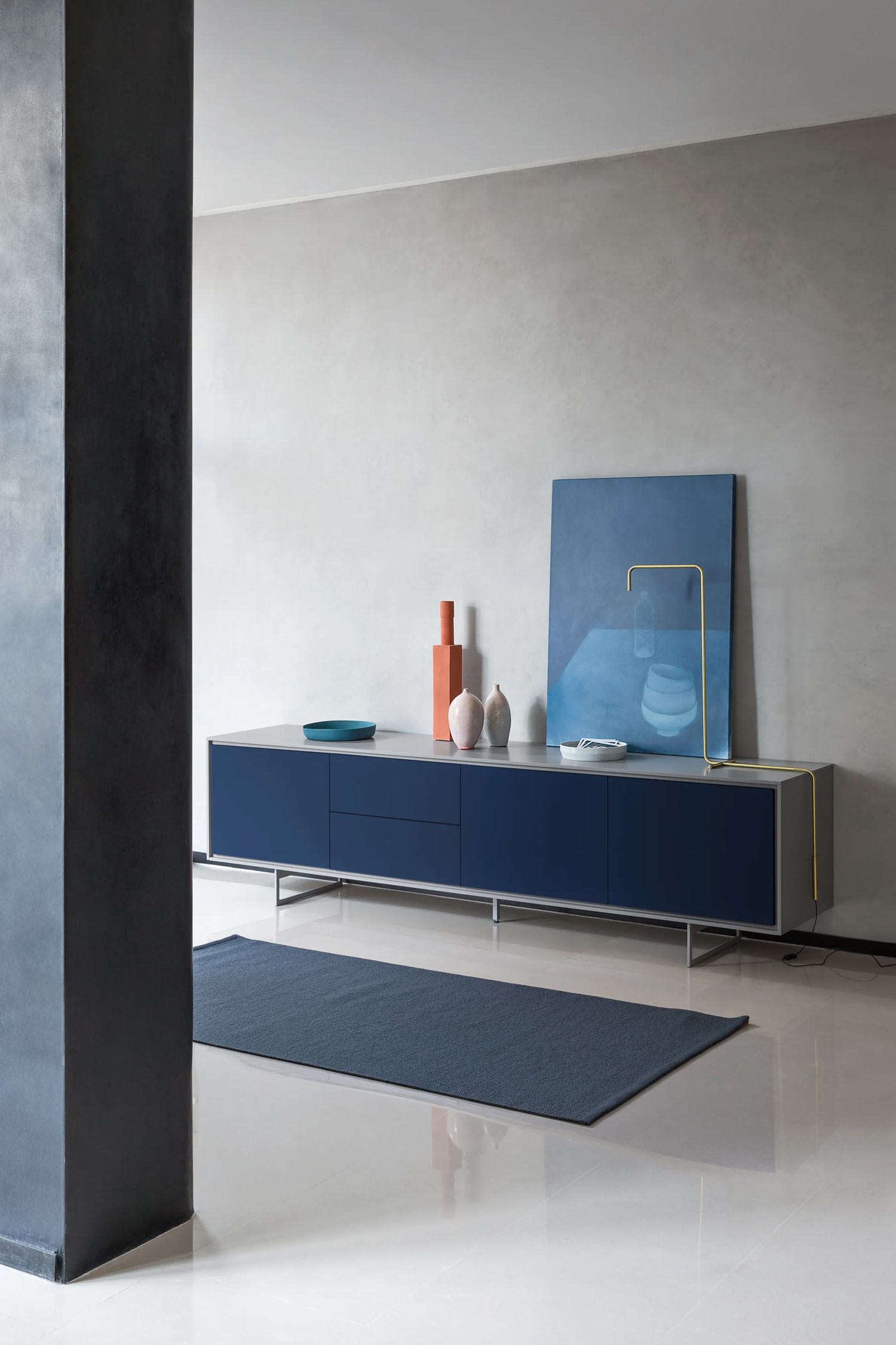 A modern living space featuring a long, dark blue and gray sideboard with two drawers and two cabinets. On top of the sideboard are various decorative items, including two tall orange vases, two light pink vases, a turquoise bowl, a white bowl, a blue abstract painting, and a unique golden-colored lamp. A dark blue rug lies on the light-colored floor in front of the sideboard. A dark gray pillar is visible on the left side of the image