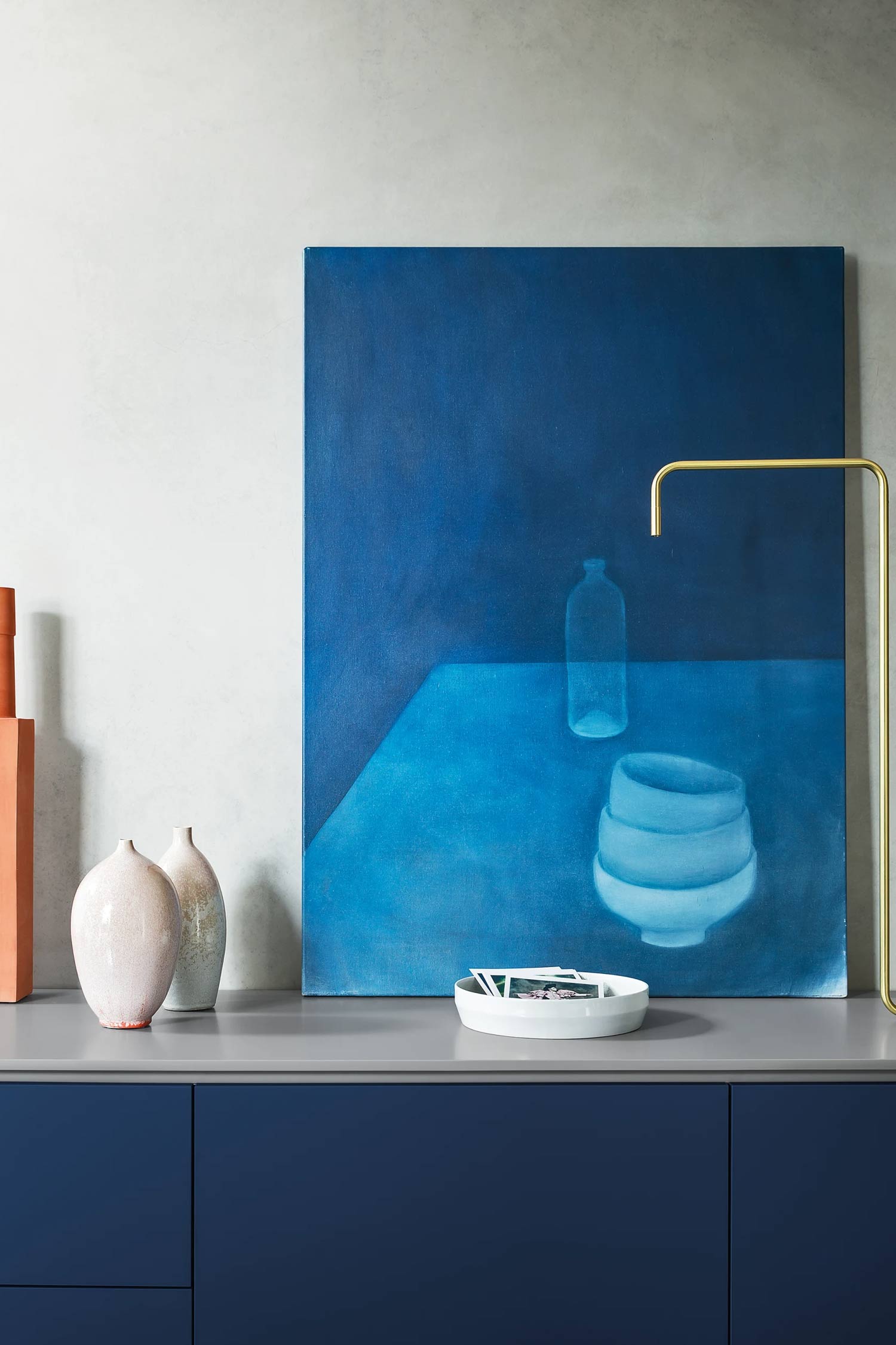 A close-up of a dark blue and gray sideboard displaying various decorative items. A large, abstract blue painting of a bottle and bowl leans against a light gray wall. In front of the painting are two light-colored vases, a small white dish with black and white photos, and a gold-colored, minimalist lamp. Part of an orange decorative object is visible on the far left.