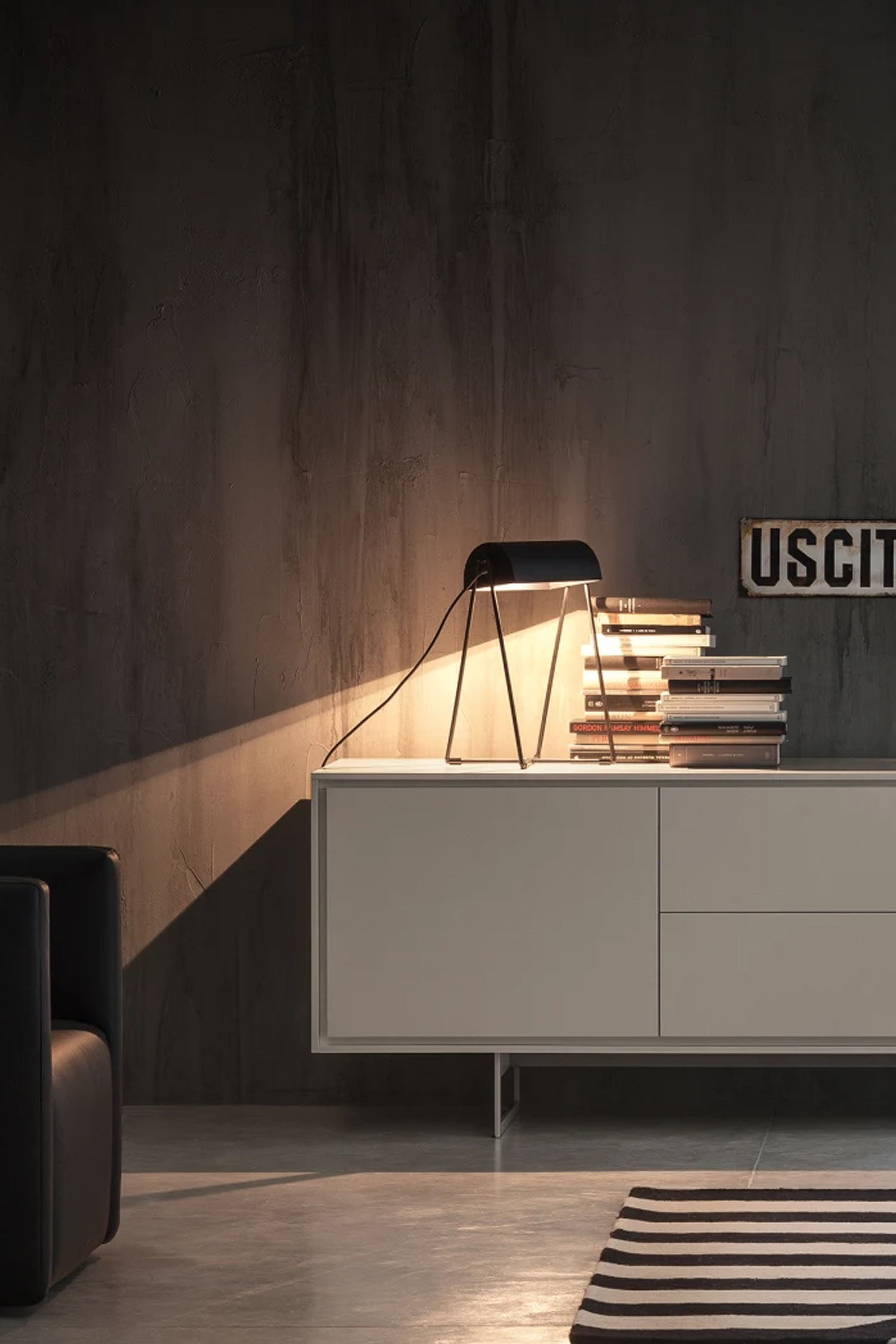 A light-colored sideboard in a dark, textured room, with a black lamp illuminating a stack of books and a small sign that says "USCITA."