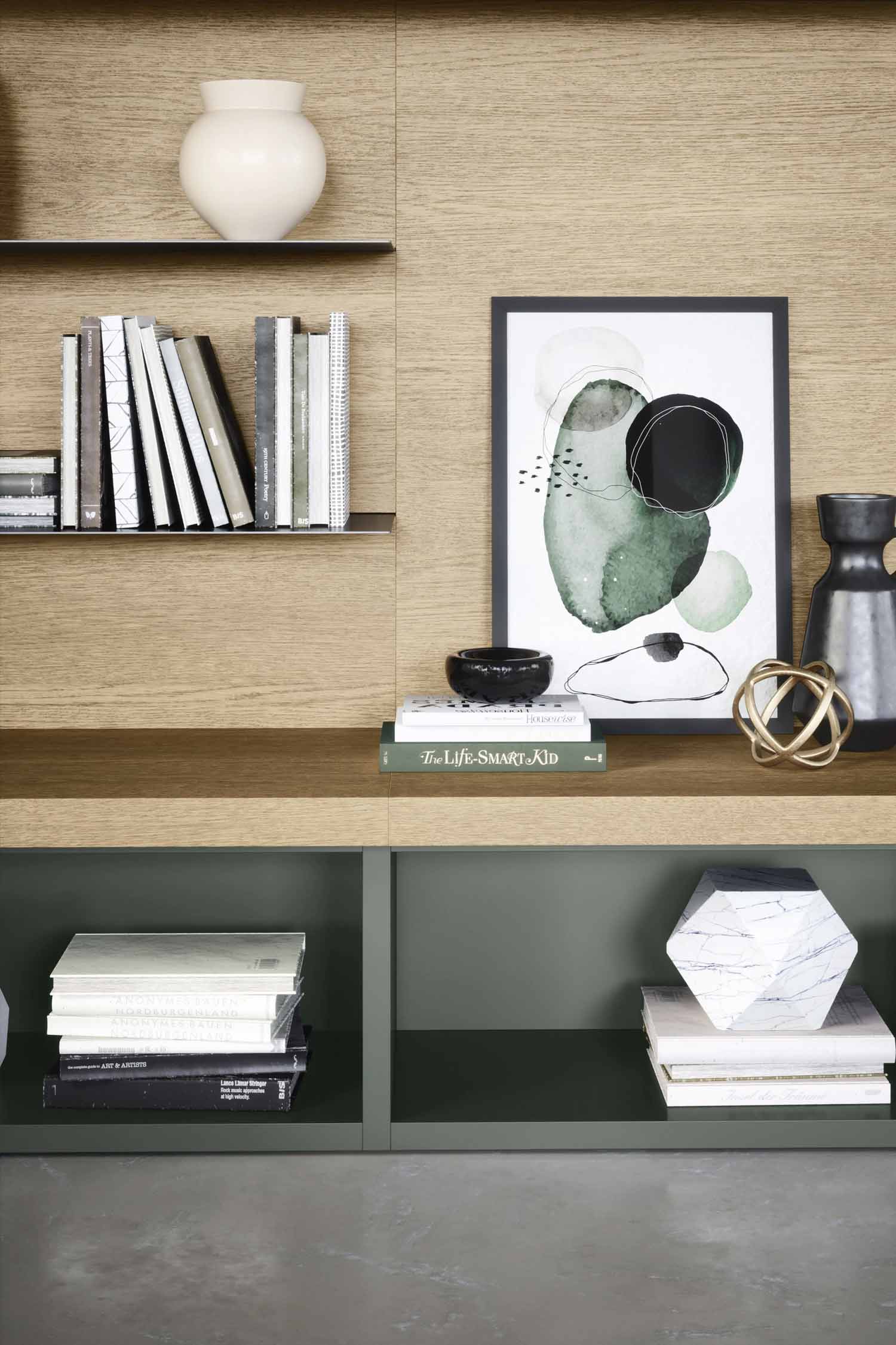 A close-up shot of a modern shelf unit. The top section is light-colored wood veneer with thin black metal shelves, holding a stack of books and a white vase. The middle section is a solid wood shelf with an abstract framed artwork in shades of green and black, a small black bowl, and a gold geometric sculpture. The bottom section is a dark green cabinet with a stack of books and a white geometric object.