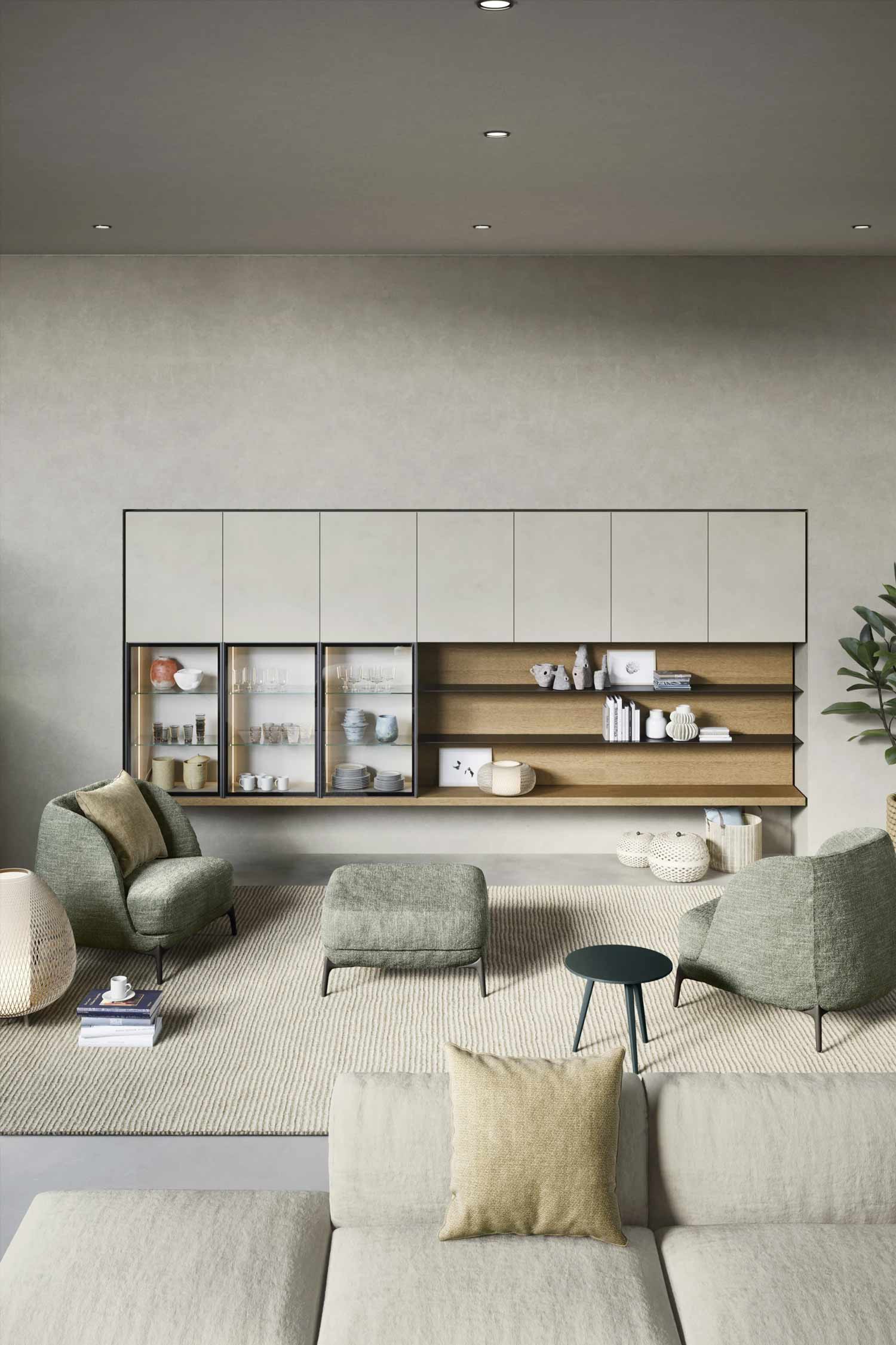 A wide-angle interior shot of a minimalist living room. A long, two-toned media unit is mounted on a textured gray wall. The top section consists of a row of light gray cabinets, while the bottom is a combination of dark-framed glass display cabinets and a light wood section with open shelves. In front of the unit, there are two matching modern armchairs and a small ottoman, all in a soft green fabric. A small round side table is placed between the chairs. The floor is covered with a light, textured rug, and a large plant is visible on the far right. A portion of a light-colored sofa is in the foreground.
