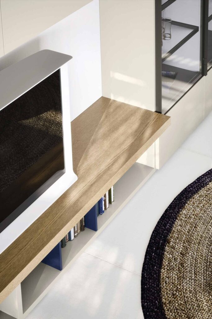 A close-up, high-angle view of a modern media console. A television with a white stand sits on a light wood shelf. The console has open storage below the shelf, where several blue and white books are visible. The unit is integrated into a larger beige wall cabinet, and a black-framed glass cabinet is visible to the right. A circular woven rug with a dark border is on the white tiled floor in the foreground.