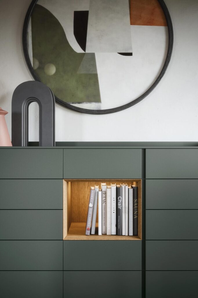 A close-up of a dark forest green dresser with six drawers, featuring a central, square, illuminated cubby lined with light wood and filled with a row of books. A modern, abstract circular art piece hangs above it.