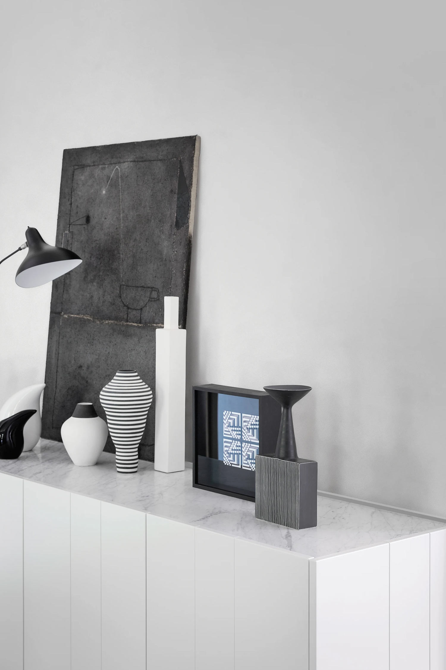 A minimalist console table with a white marble top is adorned with various decorative objects against a plain white wall. From left to right, the arrangement includes a black desk lamp, a curved white sculpture, a small white vase, a black and white striped vase, a tall white rectangular vase, a framed blue and white geometric artwork, a dark sculptural object, and a black and white striped rectangular box. A large, dark abstract painting leans against the wall behind the lamp.
